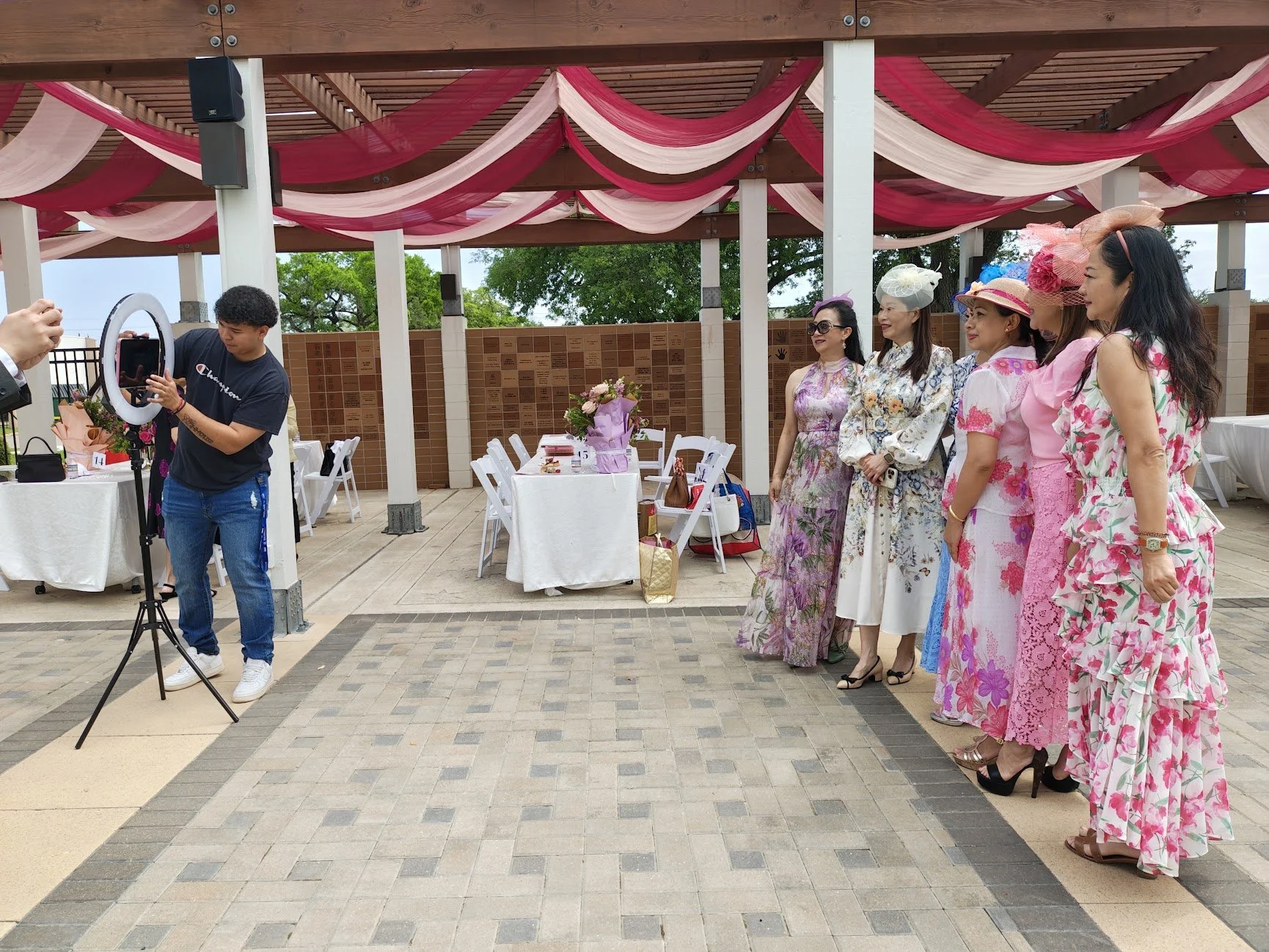 A group of women in colorful floral dresses standing together, posing for a photo at an outdoor event under a wooden pavilion decorated with pink and red fabric. A man is taking their photo using a ring light and smartphone.