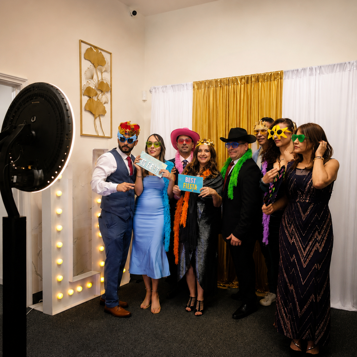 A group of eight people at a celebration or party, dressed in colorful outfits and wearing playful masks, hats, and sunglasses, standing in front of a gold and white curtain. Some are holding signs that say 'Best Fiesta' and 'Tu Fiesta'.