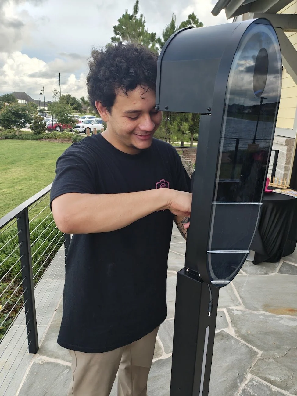 A young man with curly hair smiling and looking down while using an outdoor touchscreen kiosk on a black stand, with a grassy yard, parking lot, and cloudy sky in the background.