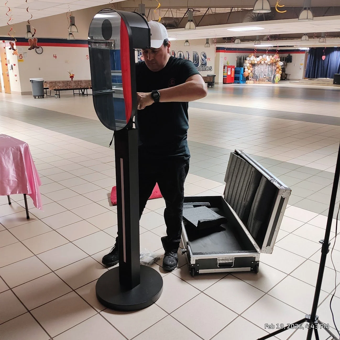 A man setting up or repairing a machine in an indoor event space, with an open equipment case on the floor nearby, decorated with balloons and banners in the background.