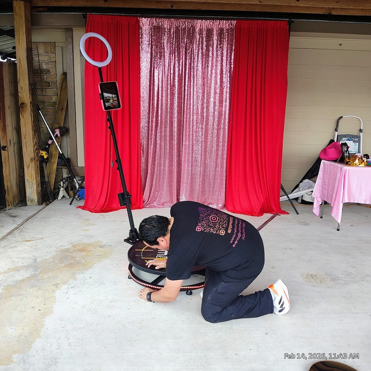 A person kneeling and leaning forward on a small punching arcade game in a room with a red and pink sequined curtain backdrop, a pink table with various items, and equipment leaning against the wall.
