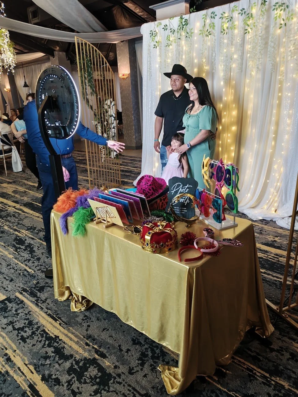 A family of three posing for a photo at a photo booth with photo props at a decorated event. The background is a white curtain with string lights, and the photo table has accessories like crowns and sunglasses.
