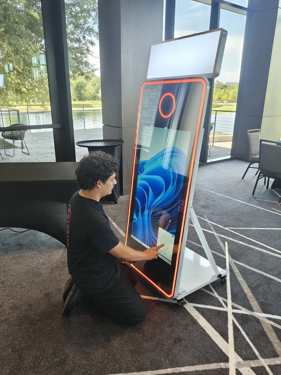 A person kneeling on the floor operating a large vertical touchscreen kiosk with a TV screen on top, near large windows showing a view outside with trees and water.