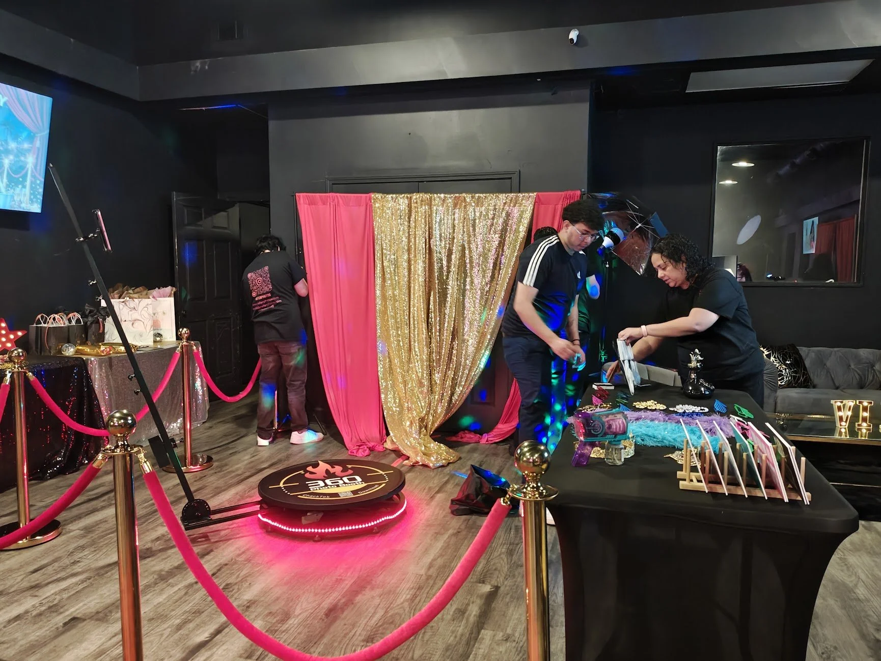 Indoor party scene with a gold sequined curtain backdrop, red curtains, a table with various items including jewelry and decorations, and a small stage with a logo reading '260 Tech' underneath red velvet ropes. Two people are setting up or preparing