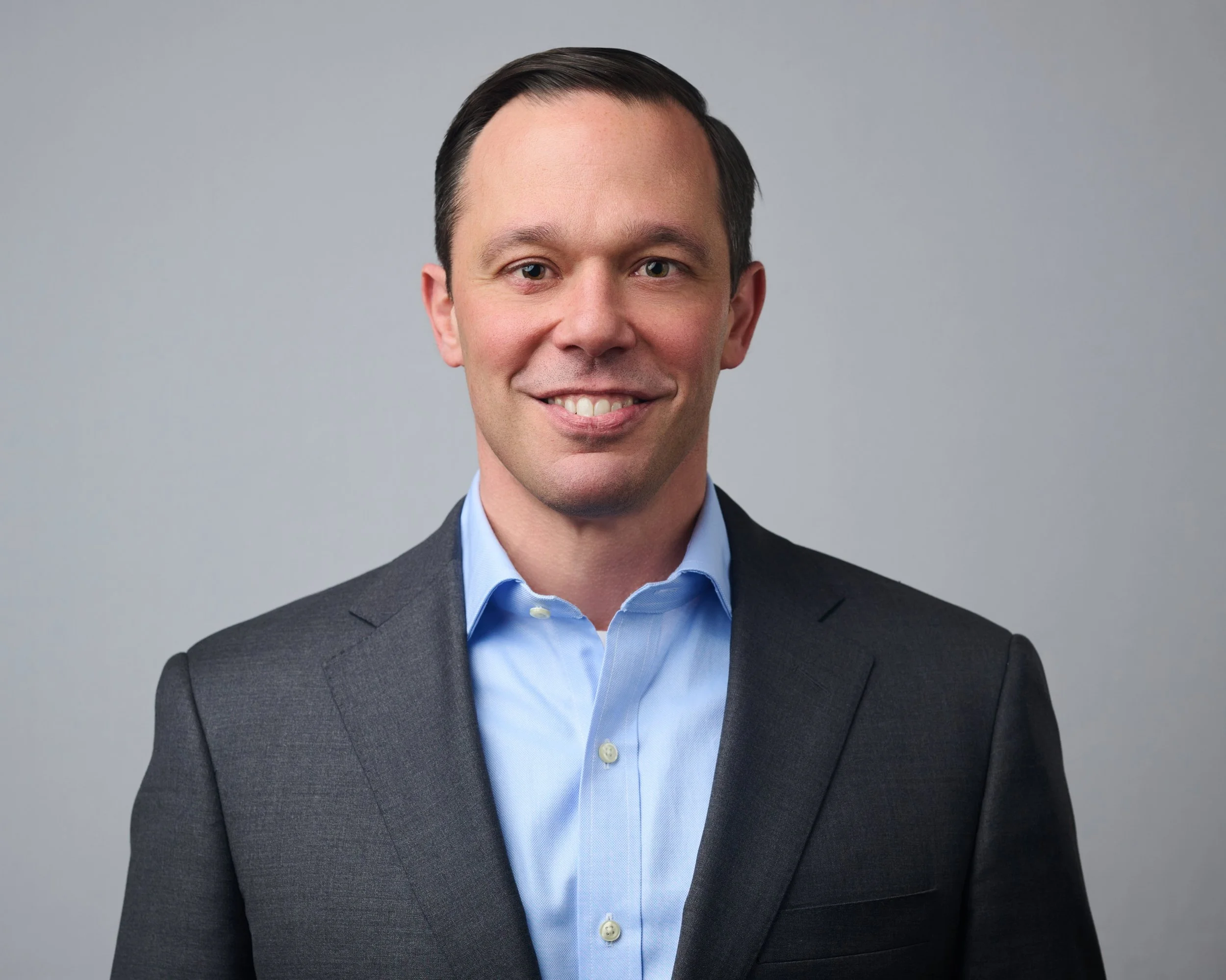 Headshot of a smiling man in a gray suit jacket and light blue dress shirt against a plain gray background.