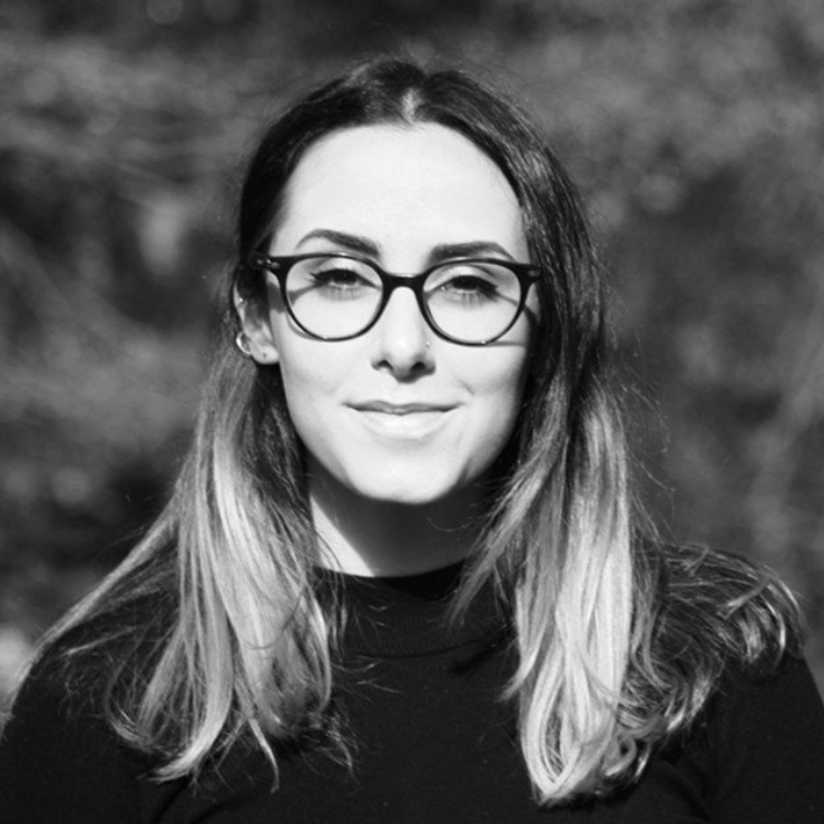 Black and white portrait of a young woman with glasses and long hair, smiling outdoors.