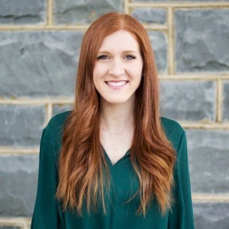 Portrait of a young woman with long red hair smiling, standing in front of a stone wall.
