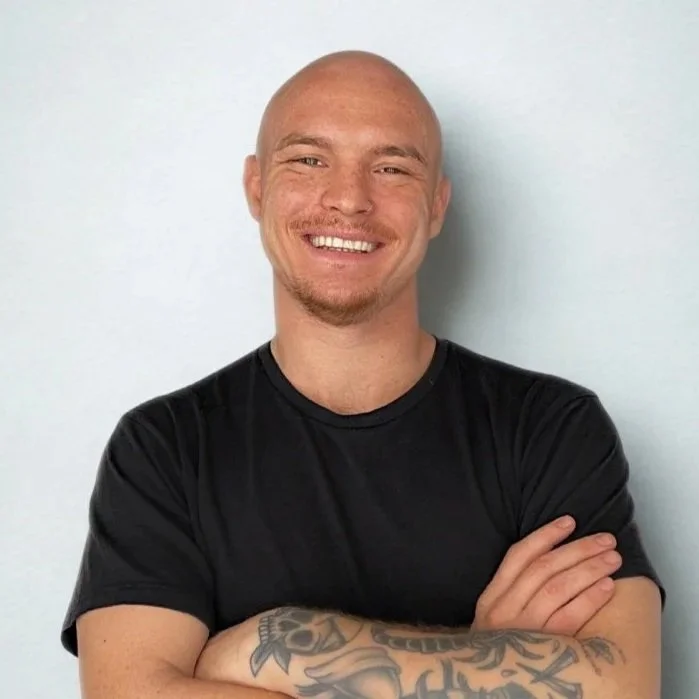 A smiling man with a shaved head, light facial hair, wearing a black T-shirt, with arms crossed, standing against a plain light background.