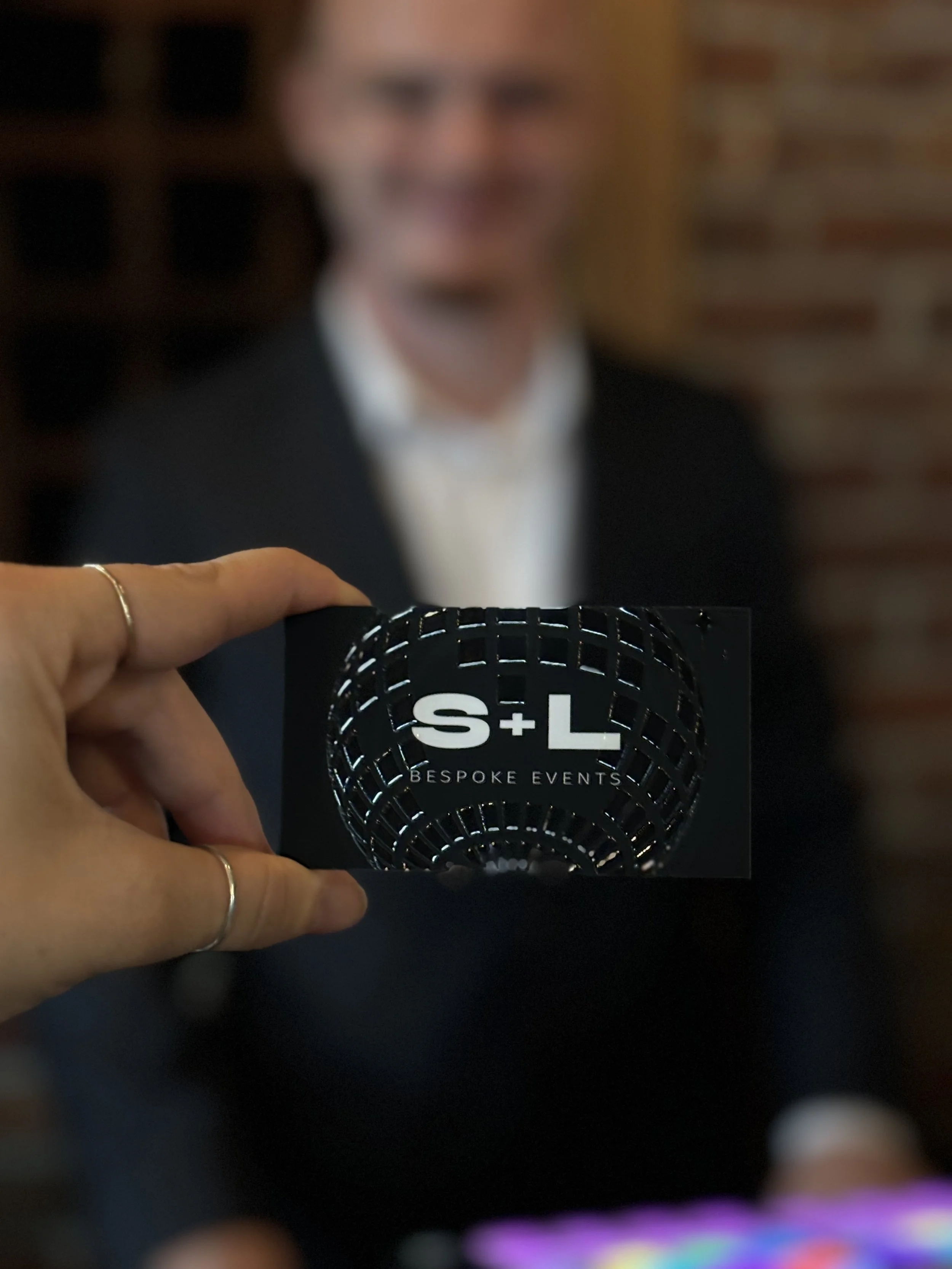 Close-up of a hand holding a business card that reads 'S+L Bespoke Events' with a blurred man in a suit in the background.