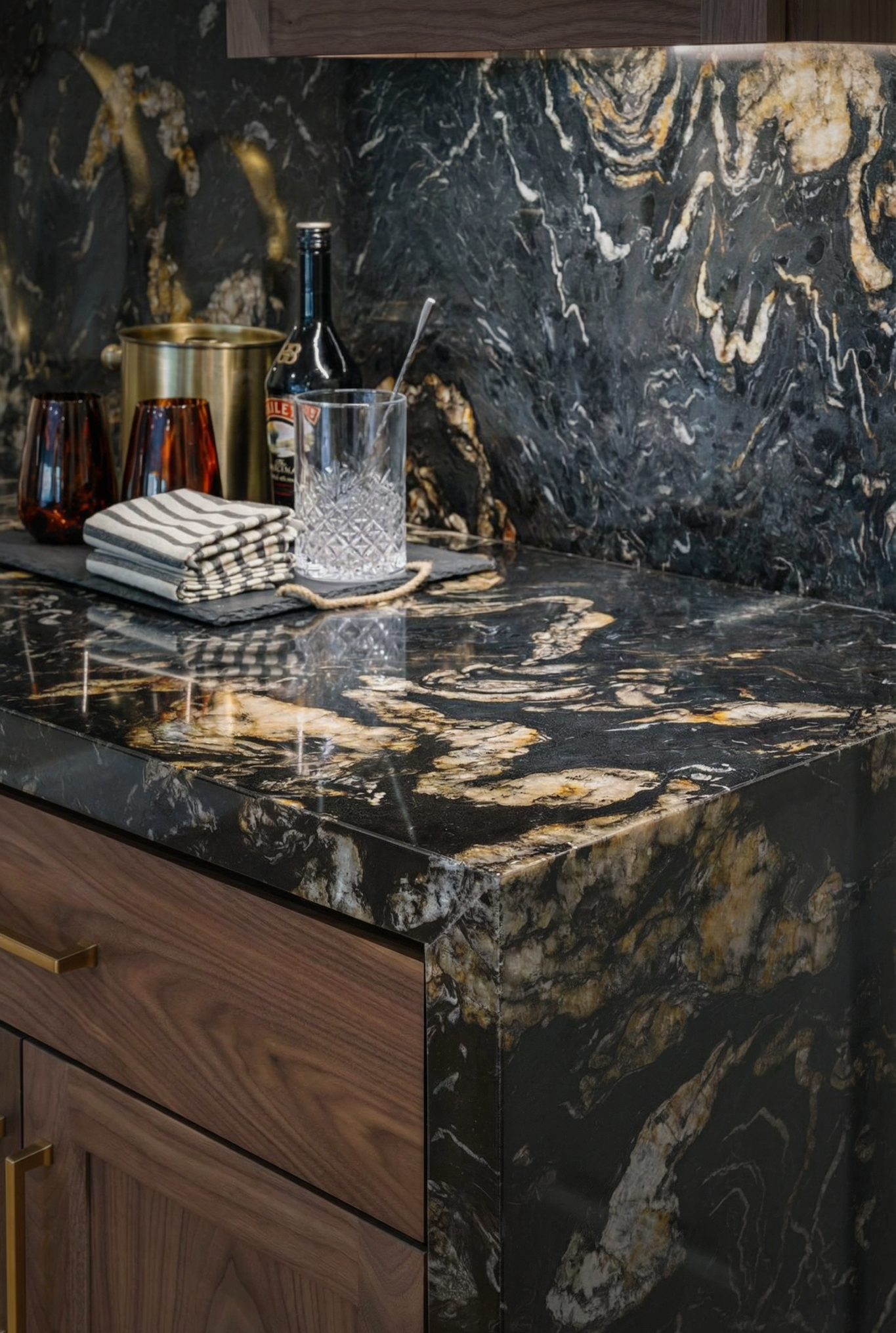 A kitchen countertop made of black marble with gold and white veins. On the counter, there are two amber-colored glasses, a folded striped cloth napkin, a glass with a stirring spoon, a bottle of tonic water, and a gold-colored ice bucket. The backgr