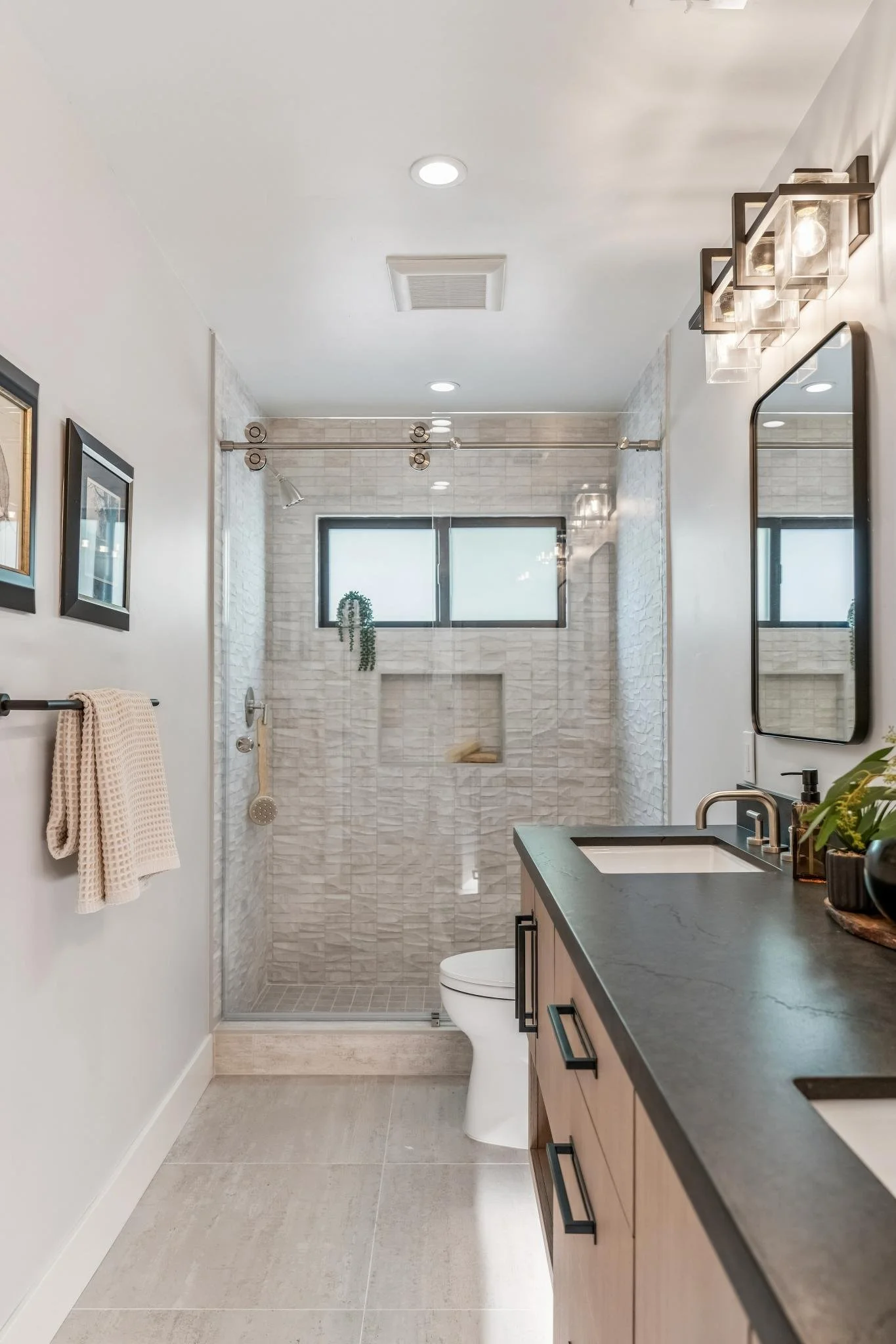 A modern bathroom with a walk-in shower with white textured tile walls and a sliding glass door, a small vanity with a black countertop and wooden cabinets, a rectangular mirror, and contemporary light fixtures. The room has beige tiled floors, a whi