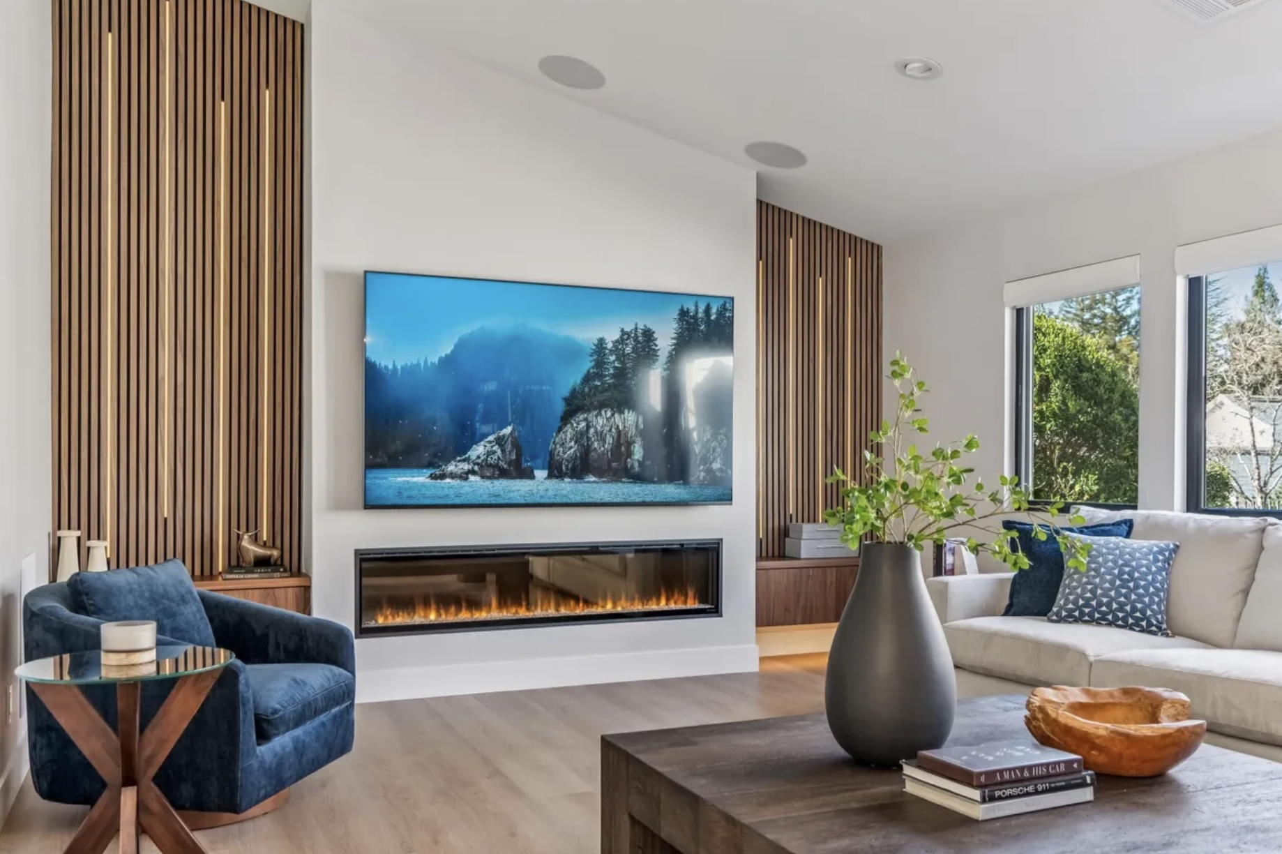 Living room with a fireplace, large wall-mounted TV showing a nature scene, a blue armchair, a white sofa with patterned pillows, potted plant on a wooden coffee table, and large windows with view of trees outside.