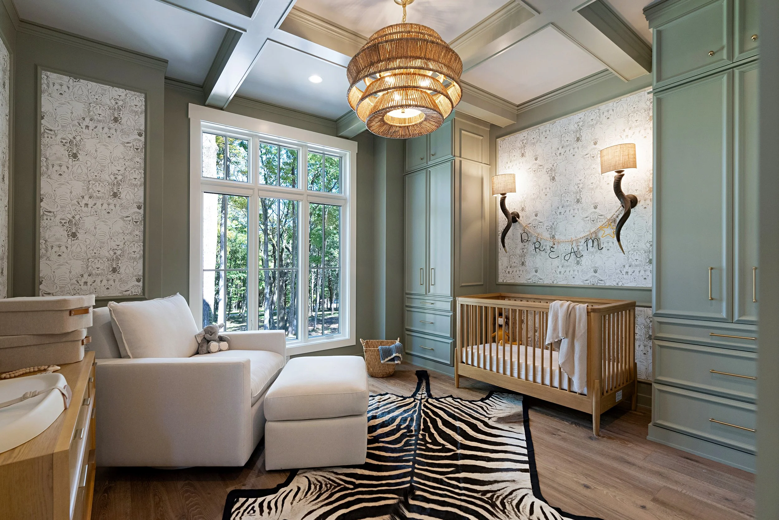 A nursery with light green walls, a large window with a view of trees, a white armchair with a teddy bear, a zebra print rug, a wooden crib, and wall-mounted lamps with animal-shaped twisted stands.