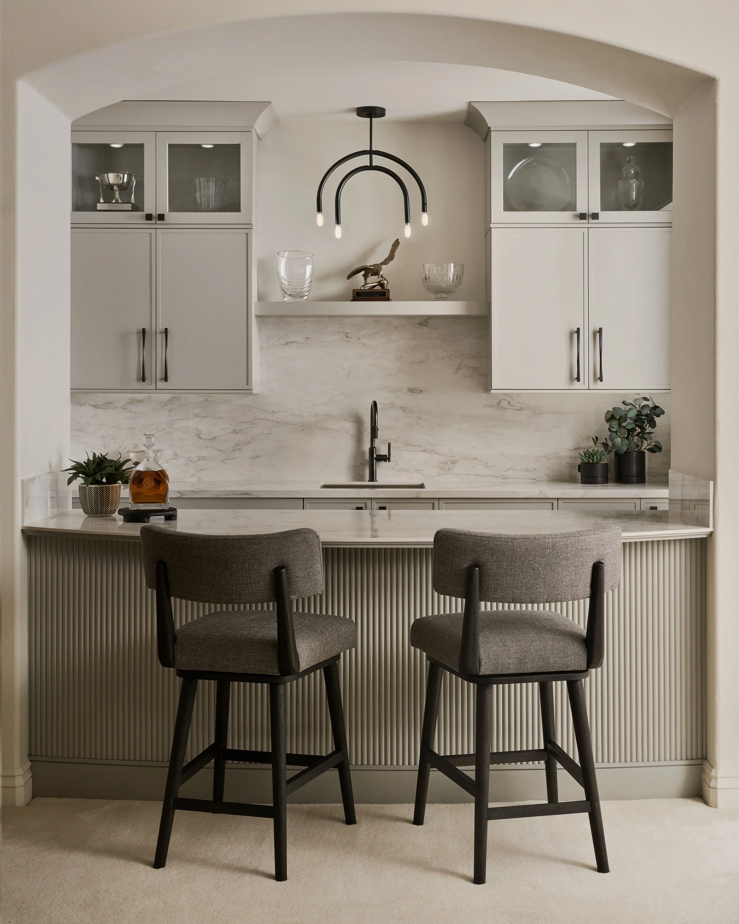 A kitchen with white cabinets, a marble countertop, and a black chandelier hanging from the ceiling. There are decorative items on a white shelf, including a glass bowl and a small trophy. There are three chairs with gray upholstery at the counter, with potted plants behind them on the right side.