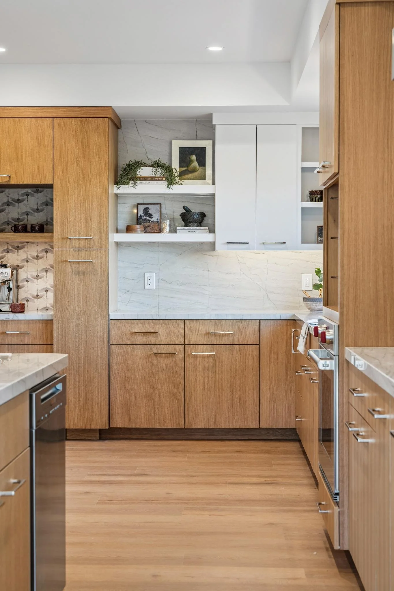 View of a modern kitchen with wooden cabinets, a marble backsplash, open shelves with decorative items, and a countertop with a sink and a coffee machine.