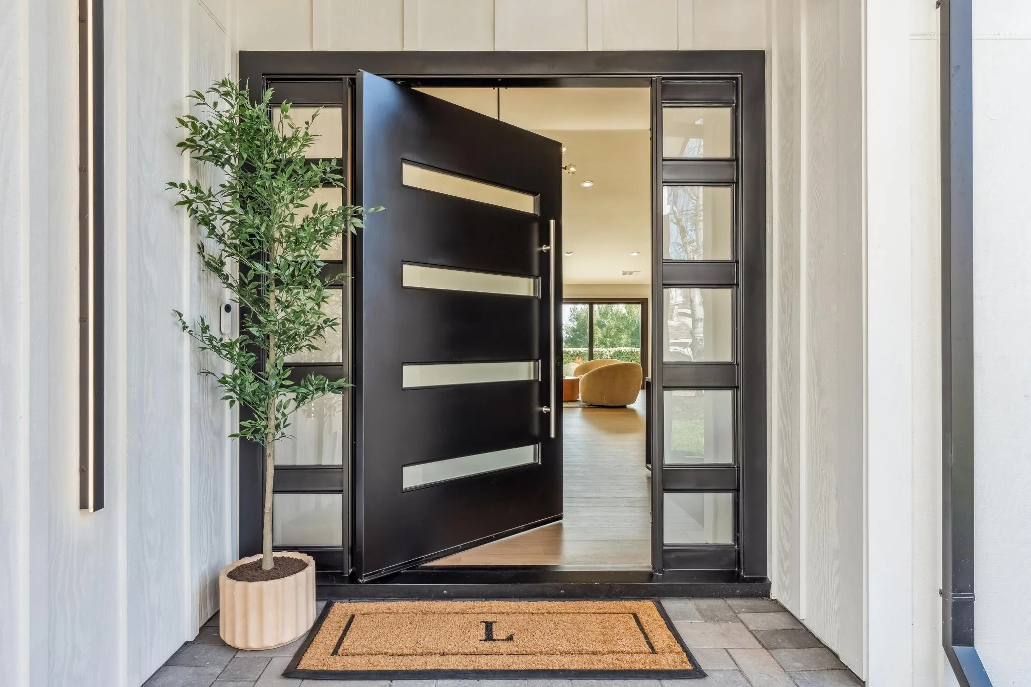 View of a modern front door entryway with a black, geometric door ajar, flanked by side glass panels. A potted green tree is positioned to the left on a mat with the letter 'L' on it, and the interior of the house with a chair and large window is vis