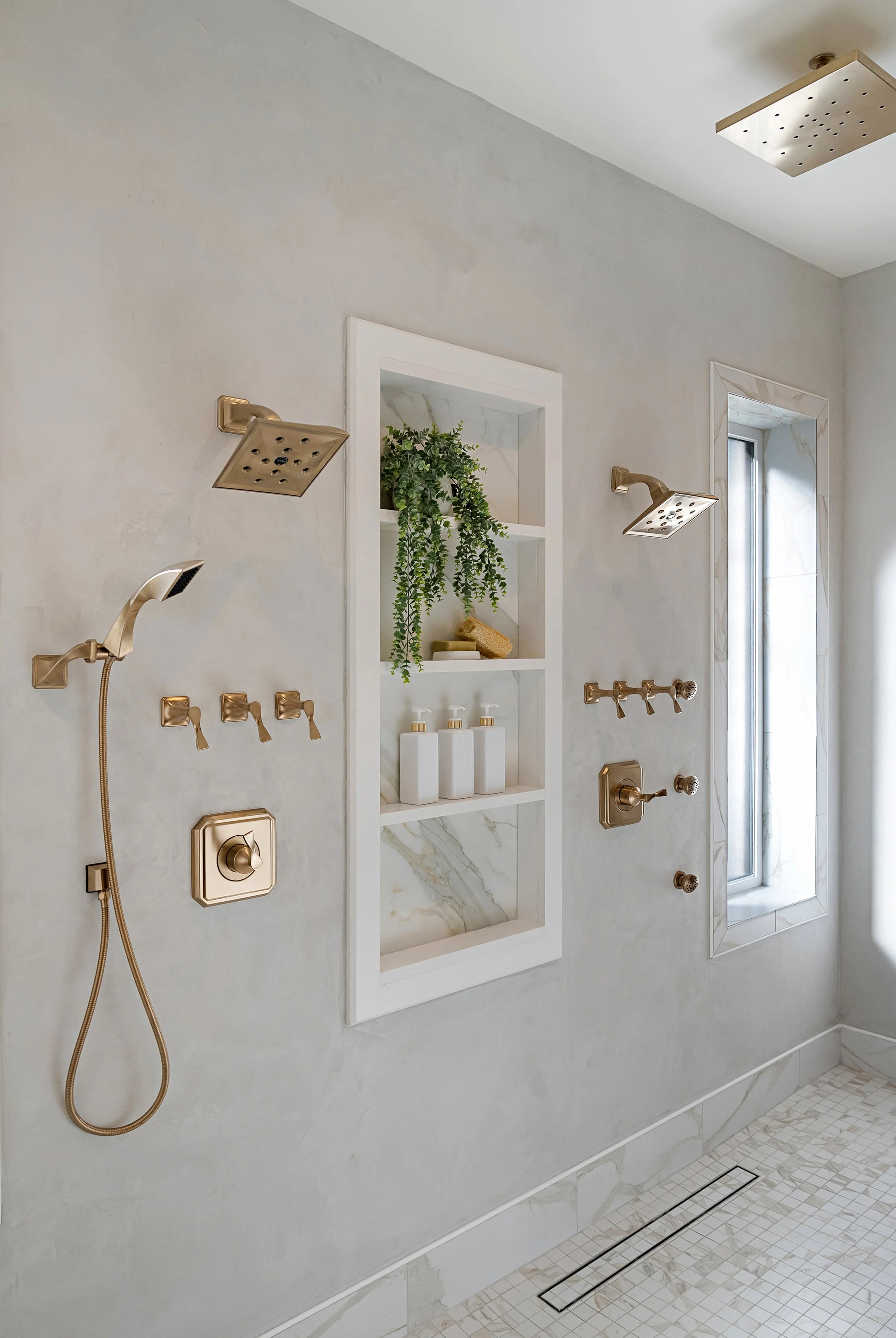 Modern bathroom with multiple shower heads, gold fixtures, and a built-in niche with a plant and soap bottles, adjacent to a window with marble trim.