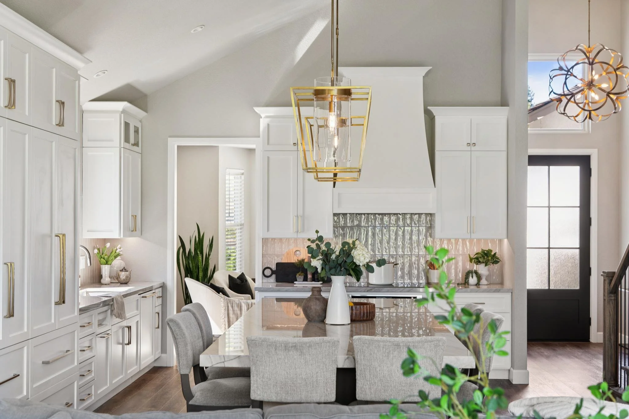 Modern kitchen and dining area with white cabinets, a marble table, and pendant lights, decorated with plants and flowers.