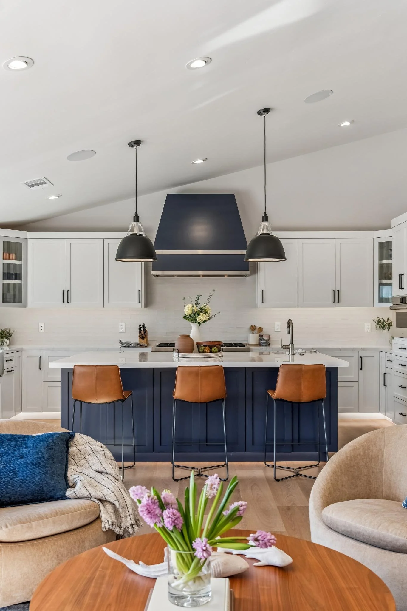 A modern kitchen with white cabinets, a navy blue range hood, black pendant lights, and a kitchen island with brown barstools. There are flowers in a white vase on the island, and cozy seating with pillows in the living area nearby.