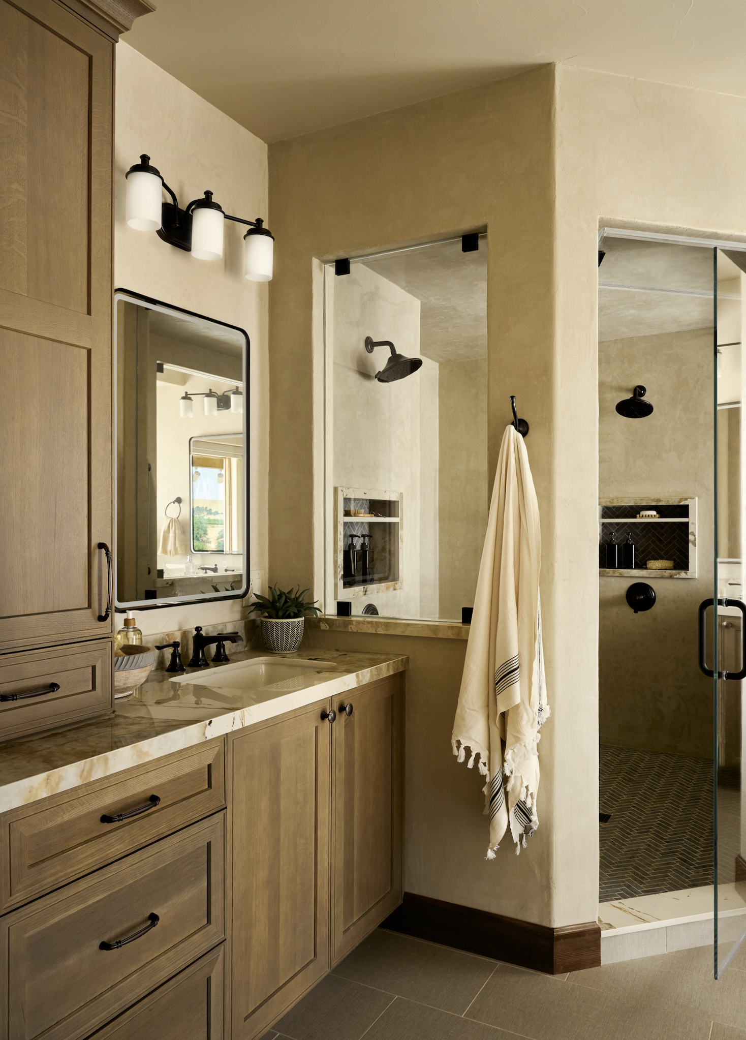 A modern bathroom with a wooden vanity, marble countertop, round undermount sink, black hardware, and wall-mounted light fixture with three frosted glass lamps. A rectangular mirror hangs above the sink, reflecting a window and a towel. Adjacent to t