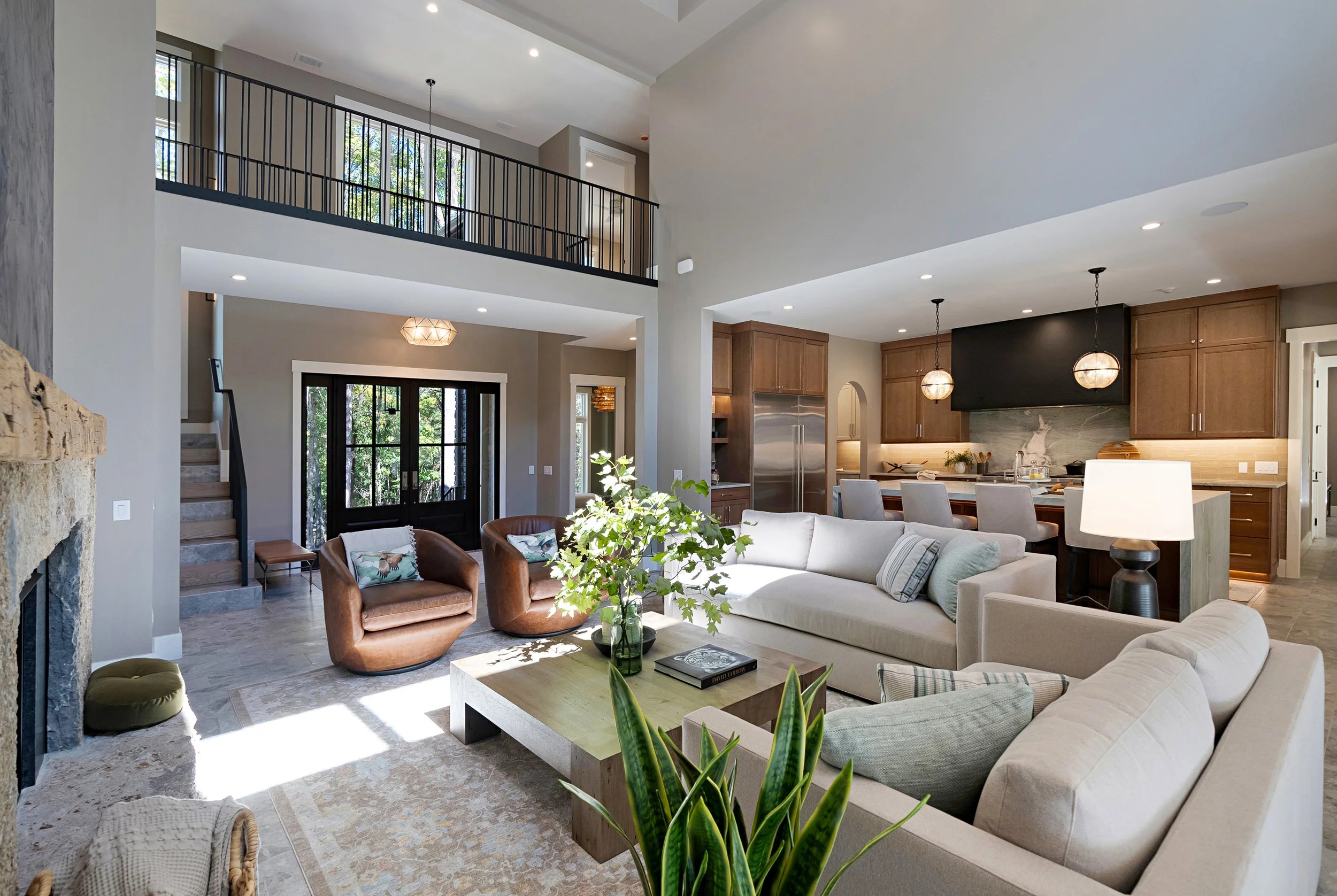 Open-concept living area with high ceilings, modern furniture, and large windows letting in natural light. The space features a beige sofa, two brown leather chairs, a wooden coffee table, and a kitchen area with wooden cabinets, pendant lighting, an