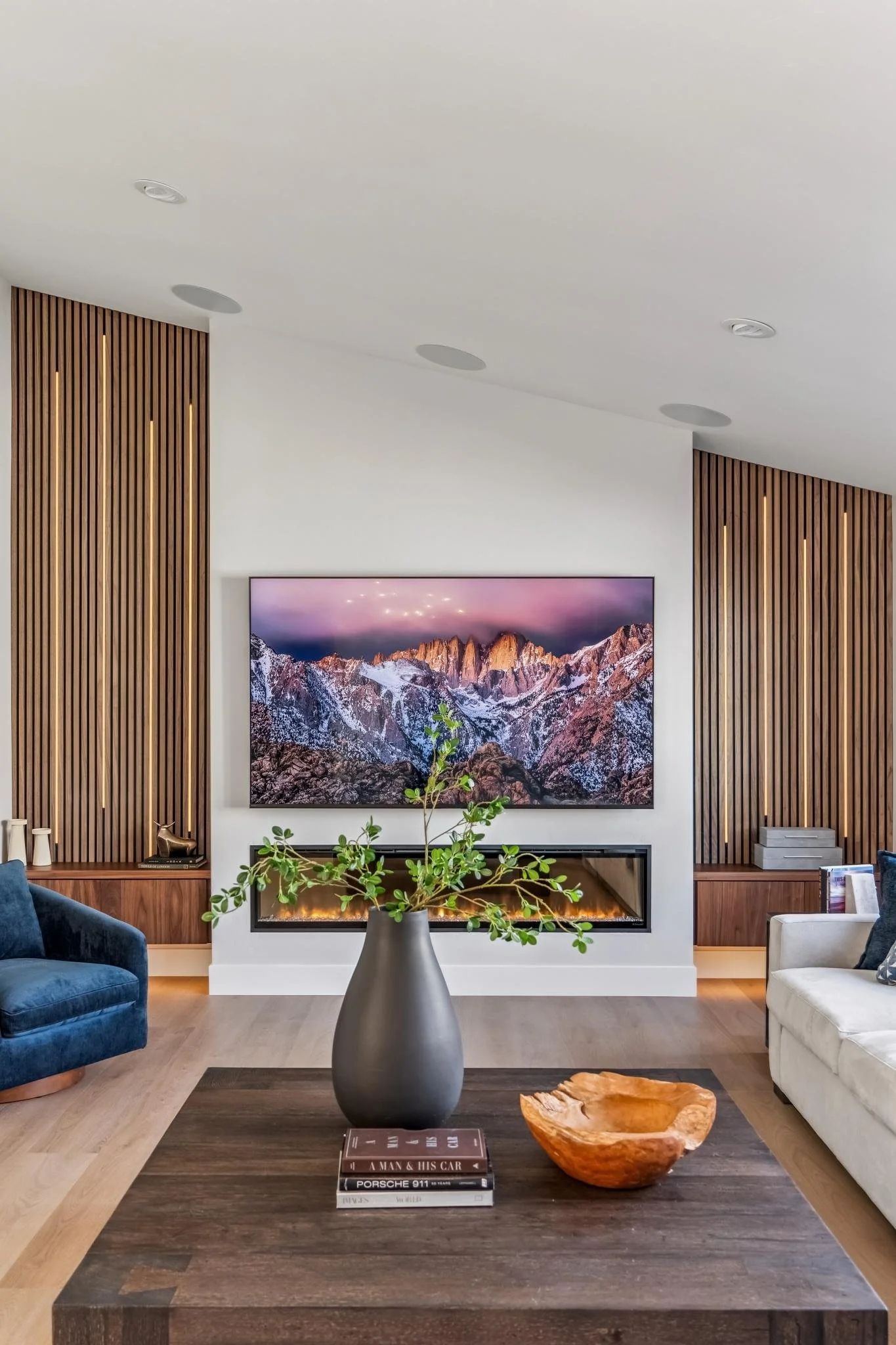 Modern living room with a wall-mounted flat-screen TV showing mountain scenery, a fireplace beneath it, wooden panel accents on the walls, a dark wood coffee table with a potted plant, books, and a wooden bowl, with two chairs, one blue velvet and on