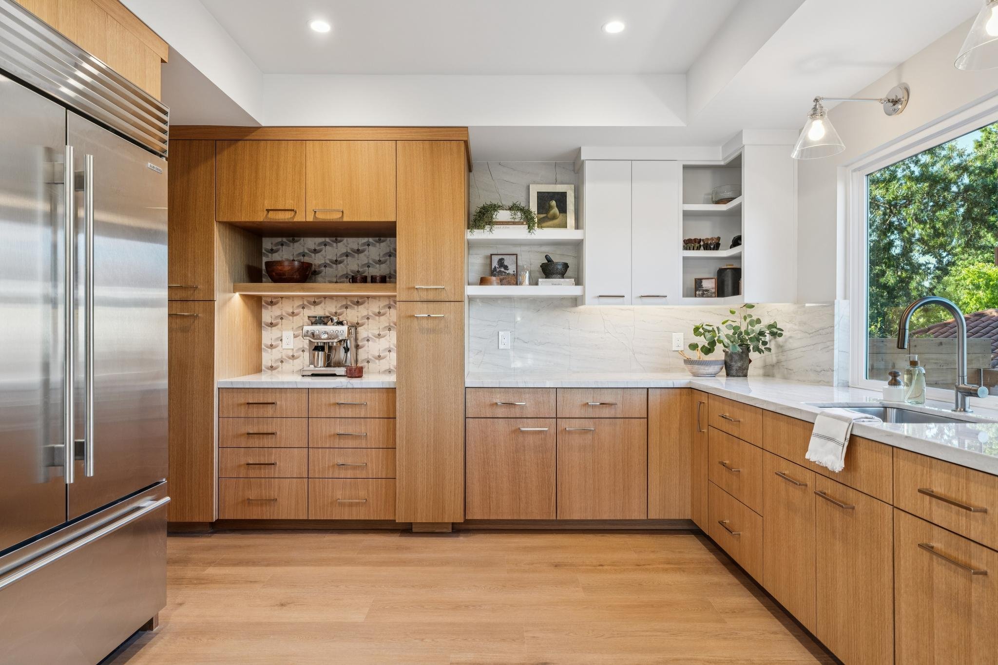 Modern kitchen with wooden cabinets, white marble countertops, and a large window with greenery outside. Contains a coffee machine, plants, and decorative items on open shelves.