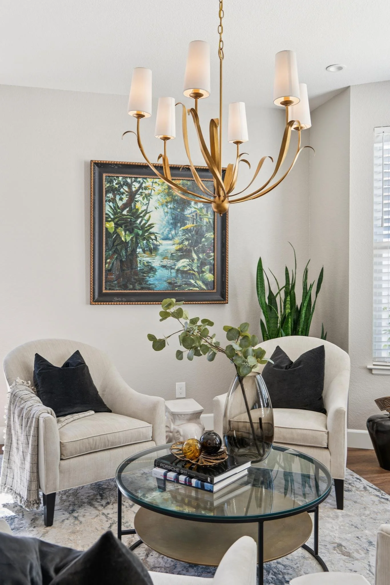 Living room scene with two white armchairs with black pillows, a glass coffee table with books and decorative objects, a large potted plant, a framed painting of a creek surrounded by trees, and a gold chandelier hanging from the ceiling.