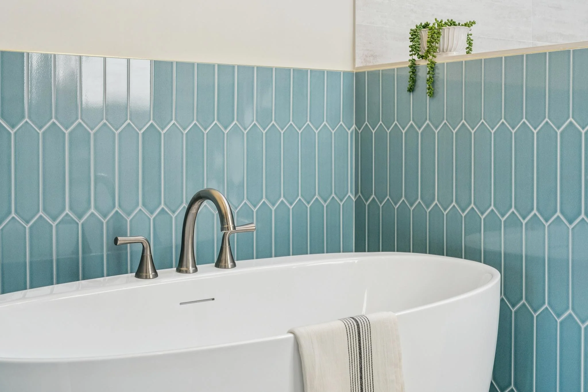Bathroom with a white bathtub, silver faucet and handles, blue tiled wall, a potted plant on a white shelf, and a folded towel hanging on the edge of the tub.