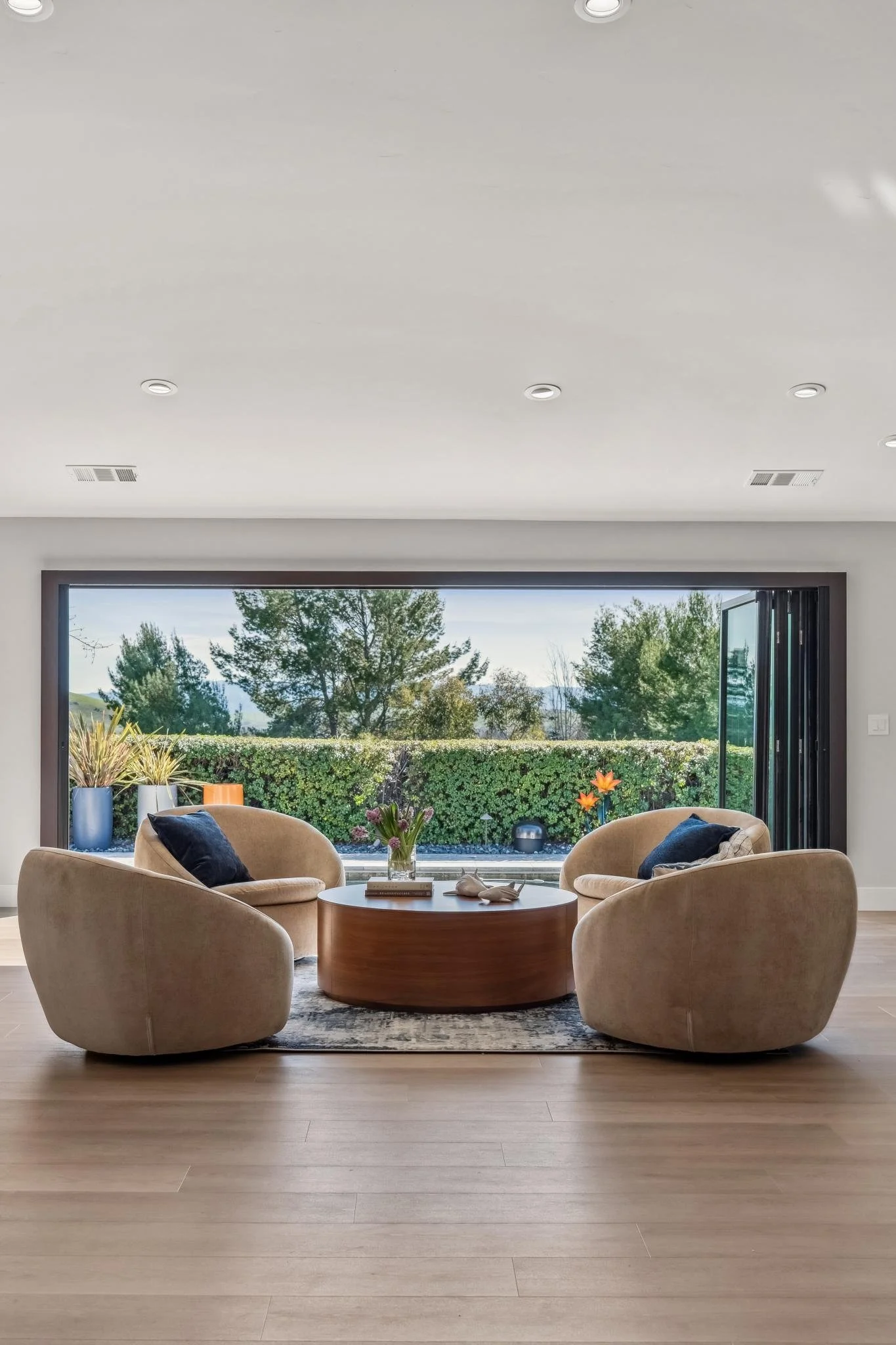 Living room with beige armchairs around a round wooden coffee table, large window with a view of trees and a hedge, decorative vases and flowers, and a patterned rug underneath.