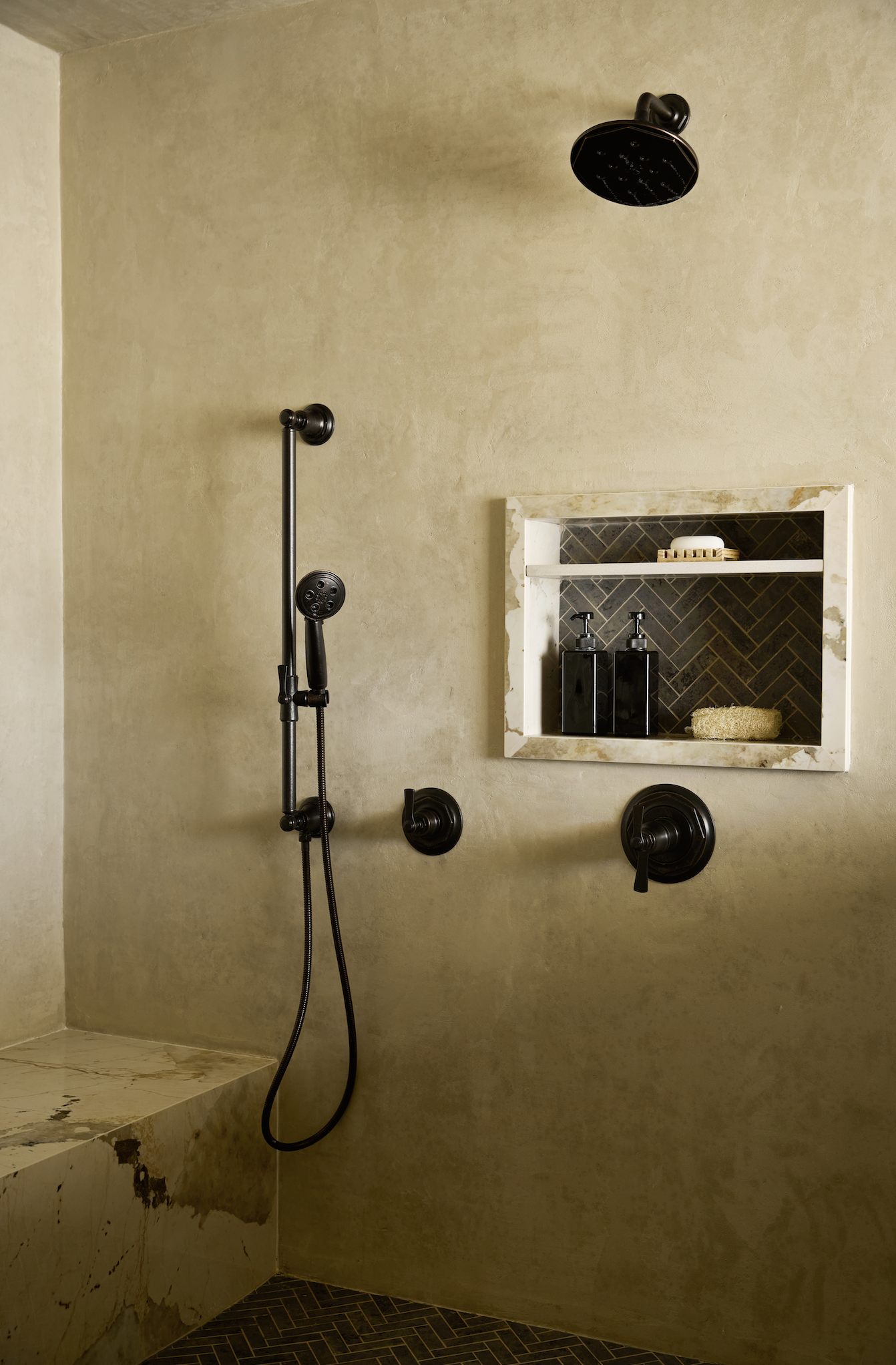 A walk-in shower with a black rainfall showerhead, black handheld shower head, and black control handles. There is a built-in shelf with soap and skincare bottles, a loofah, and a concealed bench with worn marble visible. The shower walls are texture
