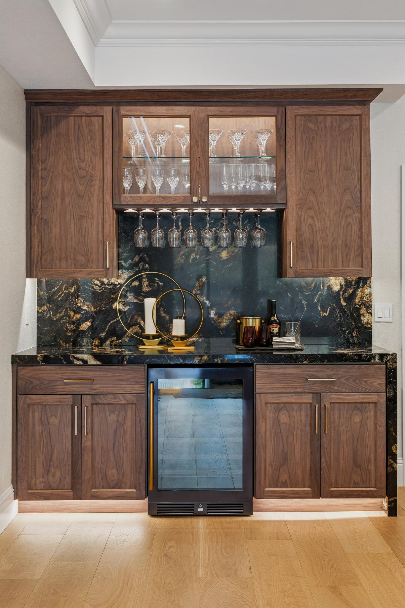 Kitchen bar area with wooden cabinetry, a black wine fridge, and a black and gold ice bucket on the countertop, with wine glasses hanging upside down and glassware displayed in glass-front cabinets.