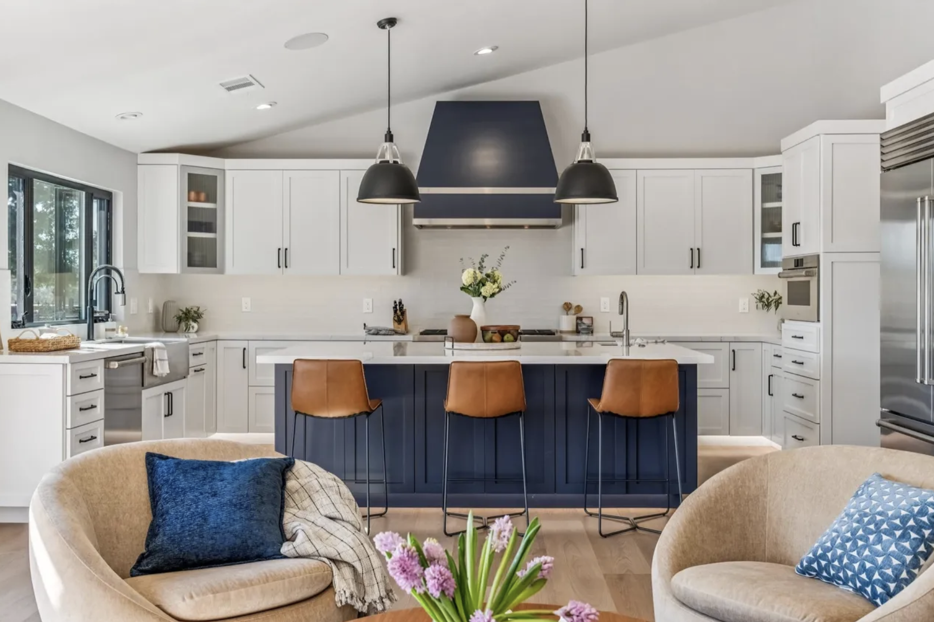 Modern kitchen with white cabinets, a navy blue kitchen island, chandelier pendant lights, and a view of living area with beige sofa and blue pillows.