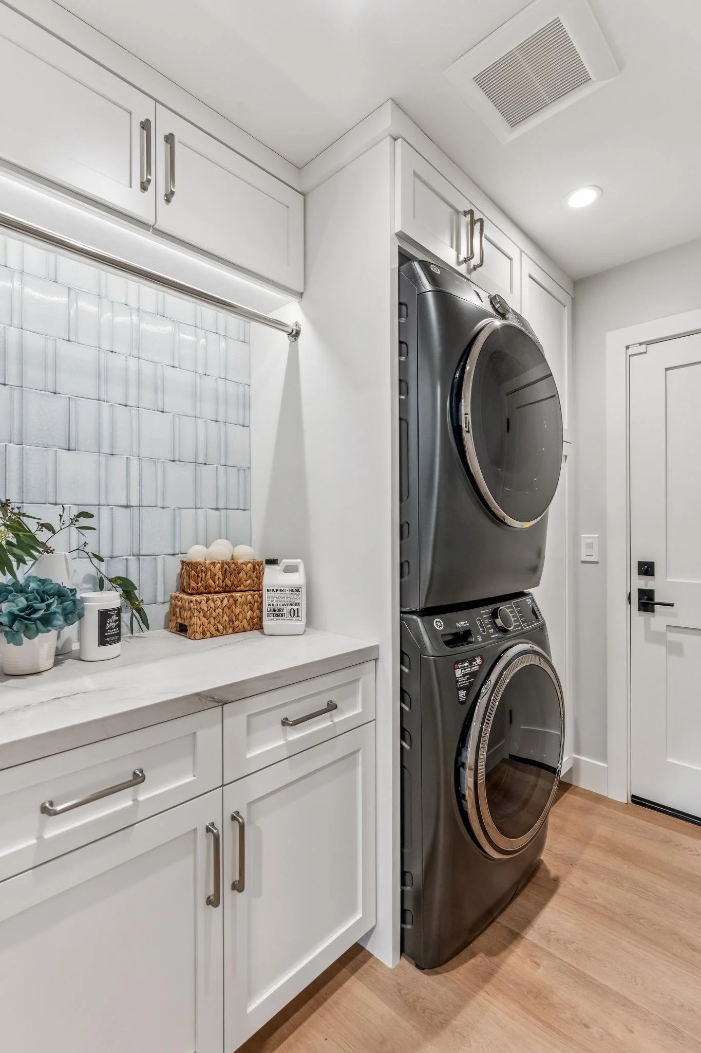 A laundry room with white cabinets, a white marble countertop, a white backsplash, a stackable washer and dryer, and decorative plants and laundry supplies on the counter.