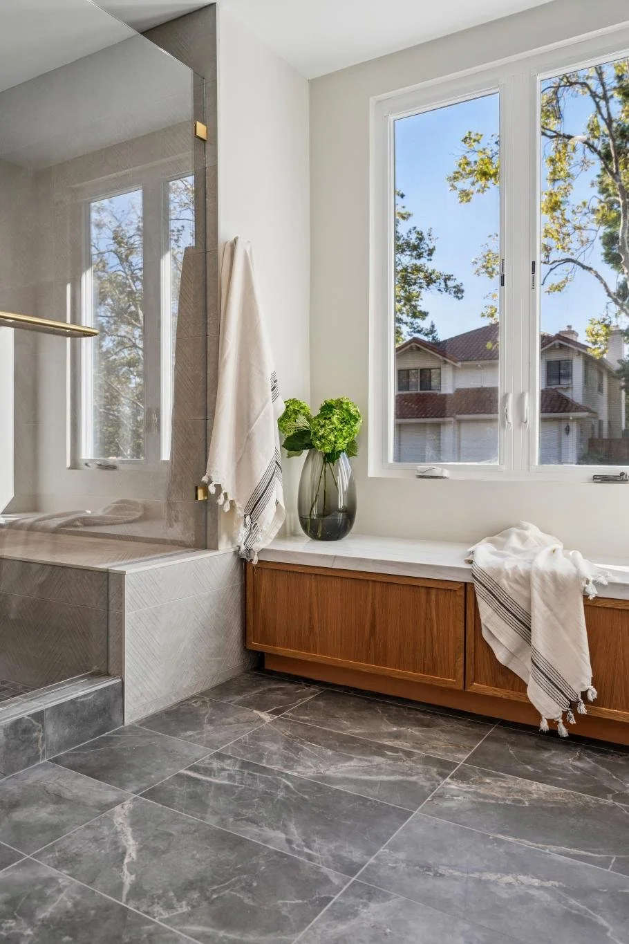A modern bathroom with large gray marble tile flooring, white walls, a built-in wooden bench, a window with a view of houses and trees, a glass-enclosed shower, a vase with green hydrangeas, and two beige towels with black stripe accents.