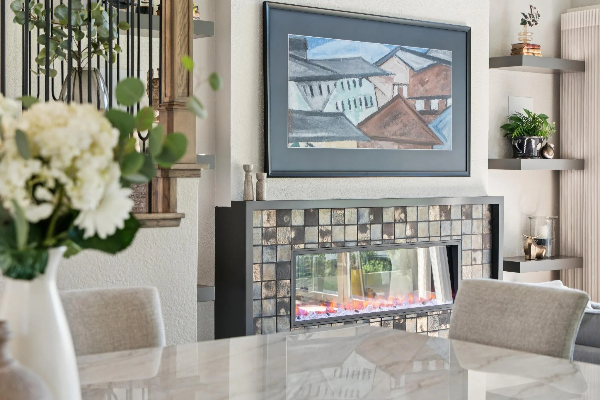 Interior of a living room featuring a fireplace with a tiled surround, a large framed painting of rooftops, floating shelves with decorative items, a potted plant, and a vase of white flowers in the foreground.