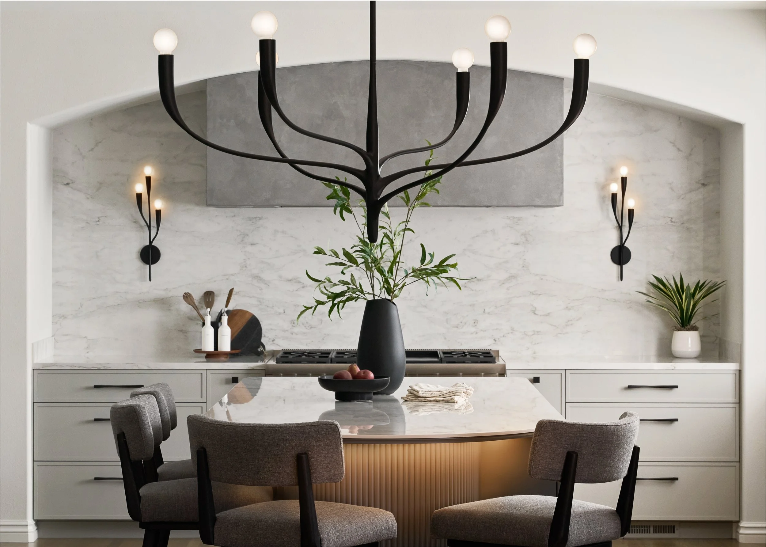 A modern kitchen with a large marble island, gray chairs, a black vase with green leaves, and stylish wall-mounted lights.