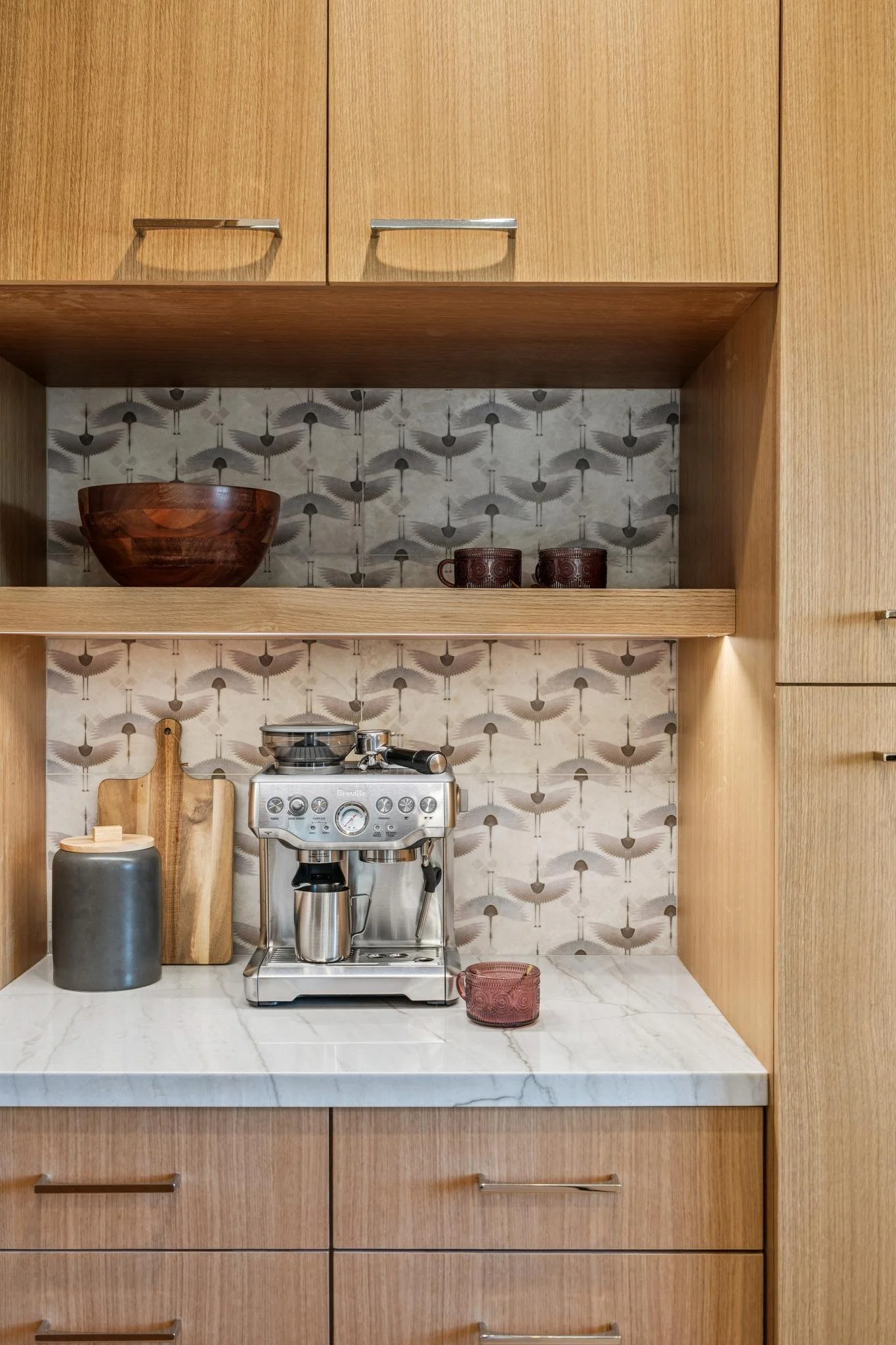 Kitchen counter with a coffee machine, two mugs, a wooden bowl, a cutting board, a jar, and a spice grinder, with wooden cabinets and patterned wallpaper in the background.