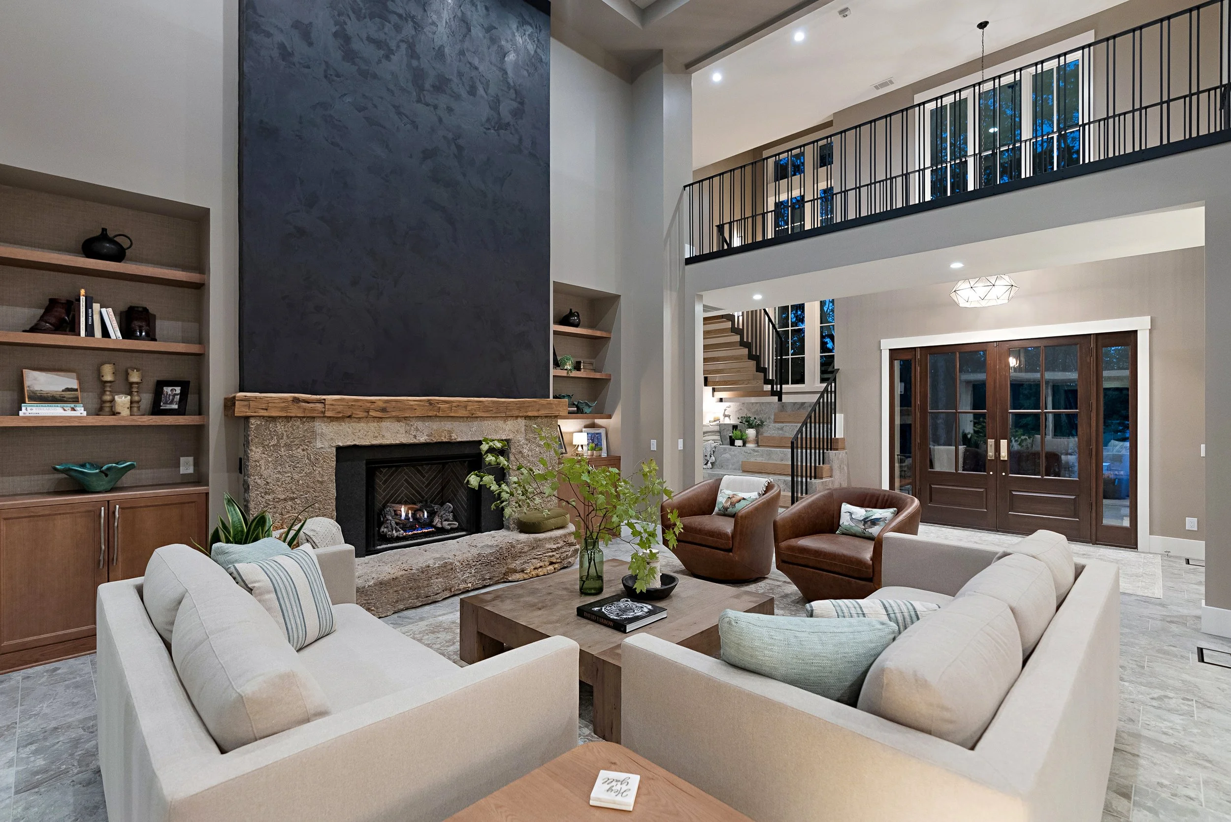 A spacious living room with a stone fireplace, built-in wooden shelves, modern staircase, large windows, and ceiling lights. Contains white and brown seating, a wooden coffee table, plants, and decorative items.