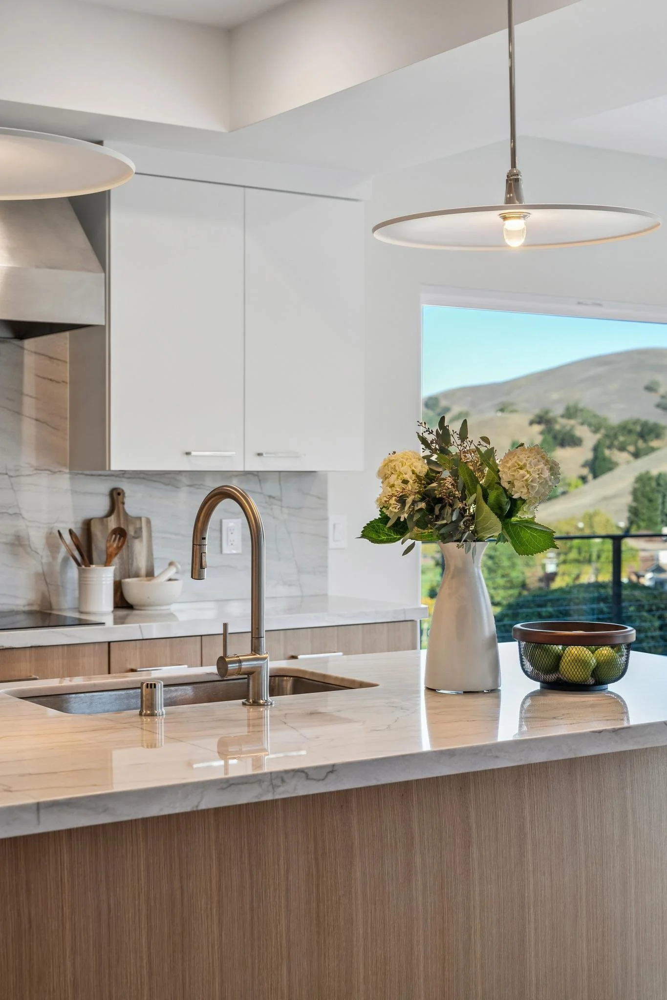 Modern kitchen with white cabinets, a marble countertop, a sink with a brass faucet, a vase with flowers, a bowl of green apples, and a scenic view of hills through the window.