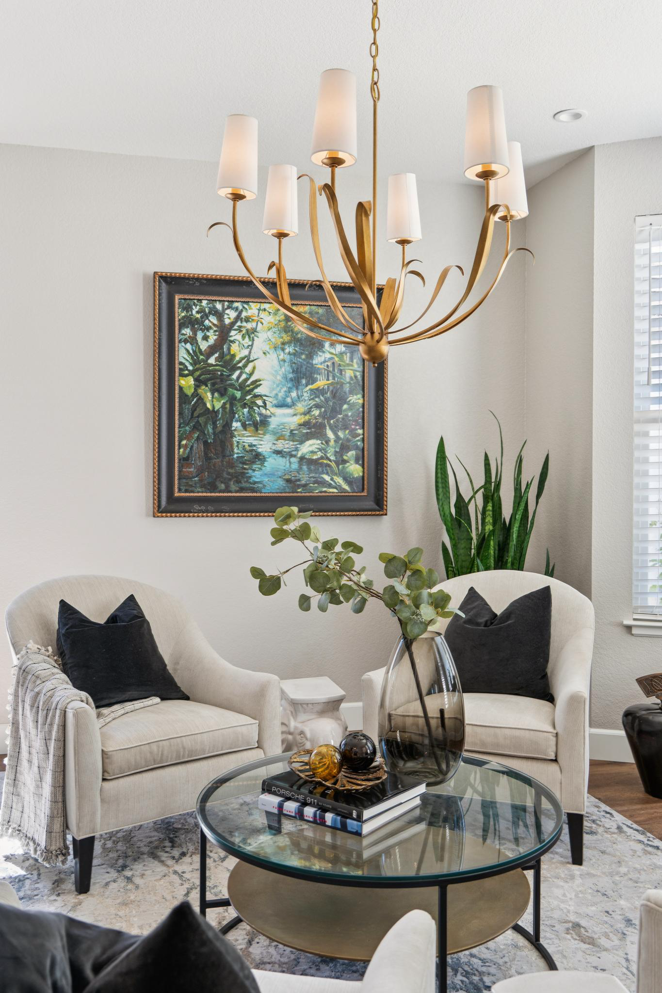 Living room with cream sofas, black pillows, and a glass coffee table with books and decorative objects, a large painting of a forest scene on the wall, a gold chandelier hanging from the ceiling, and tall houseplants near a window with blinds.