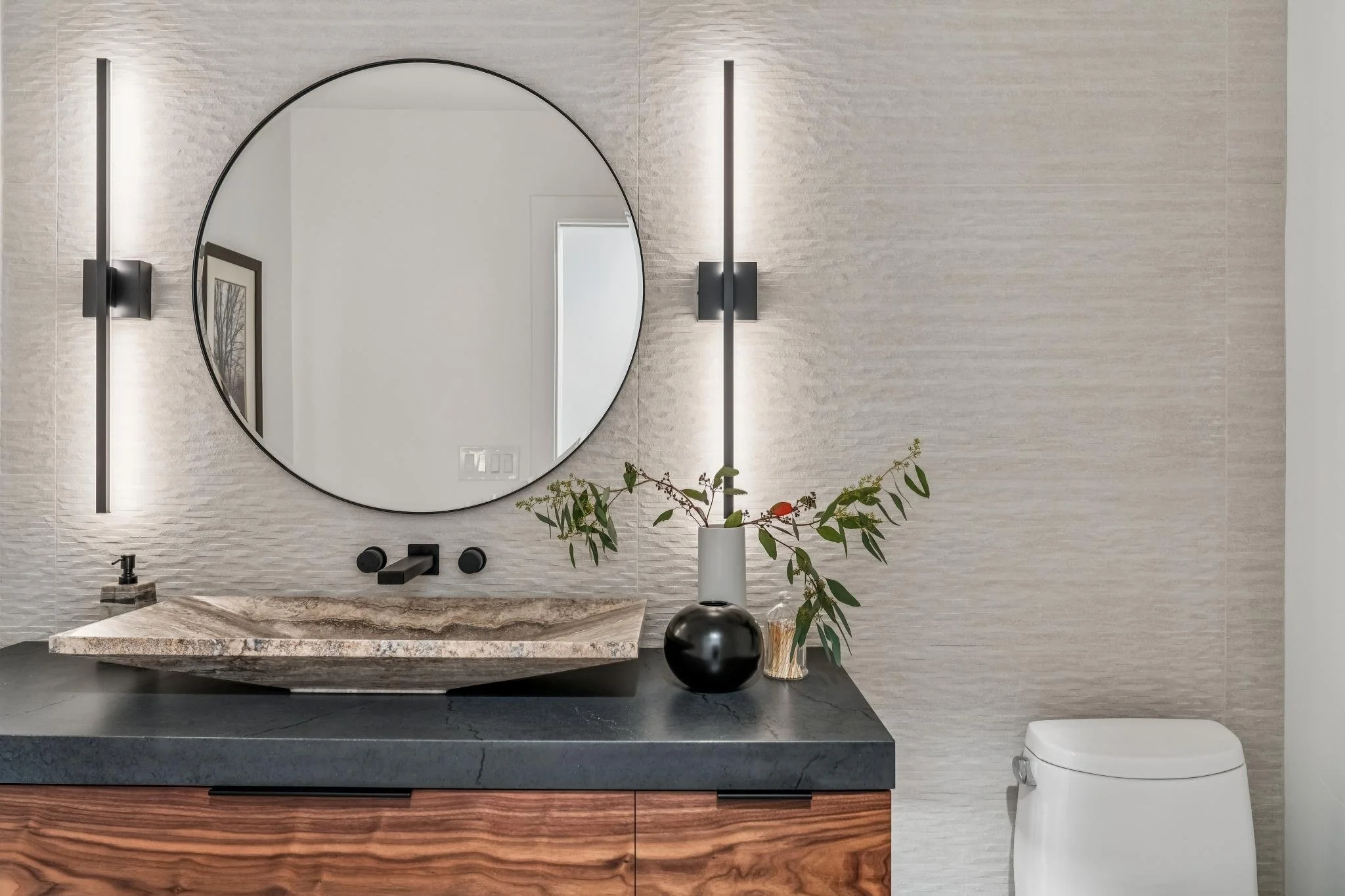 Modern bathroom vanity with a large round mirror, black fixtures, and a beige granite countertop. A vase with green leaves and a small decorative container are on the counter, with white textured tile walls and a white toilet to the right.