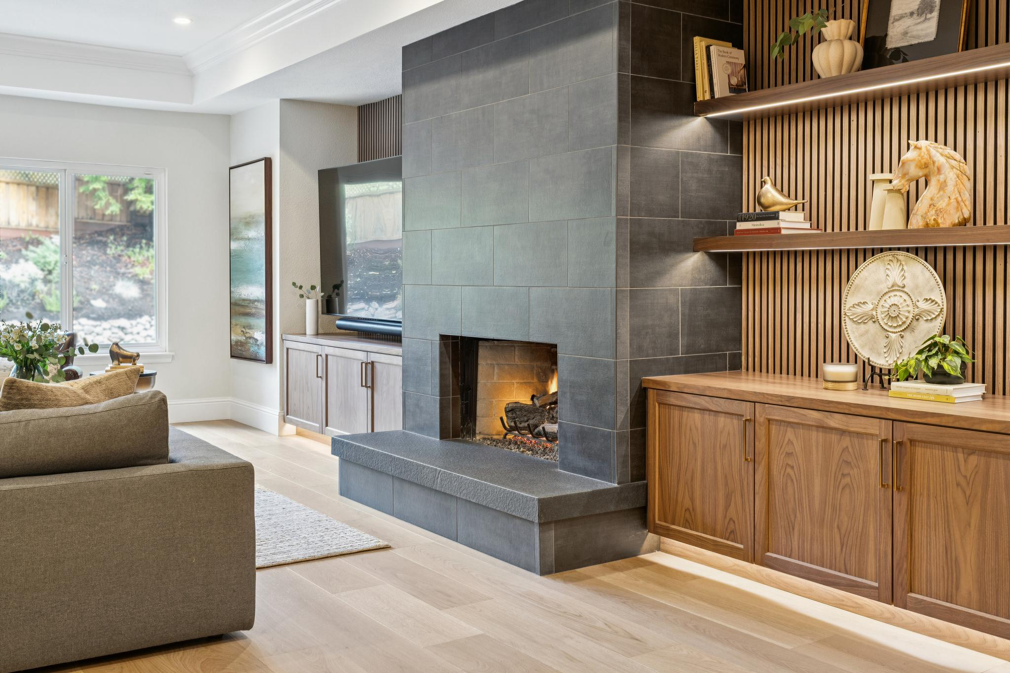 Living room with a modern gray fireplace, wooden cabinetry, and decorative items on shelves, including a horse sculpture and stacked books. Sunlight enters through large windows.