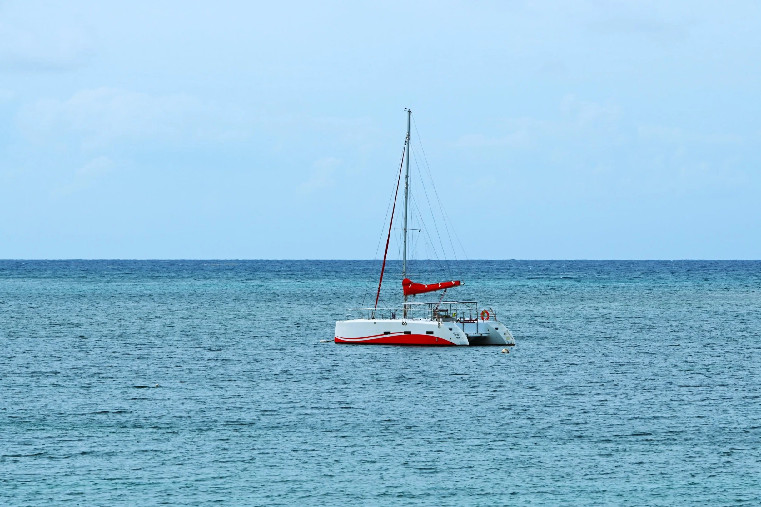 A white sailboat with a red stripe on the hull floating on the ocean, with an overcast sky in the background.