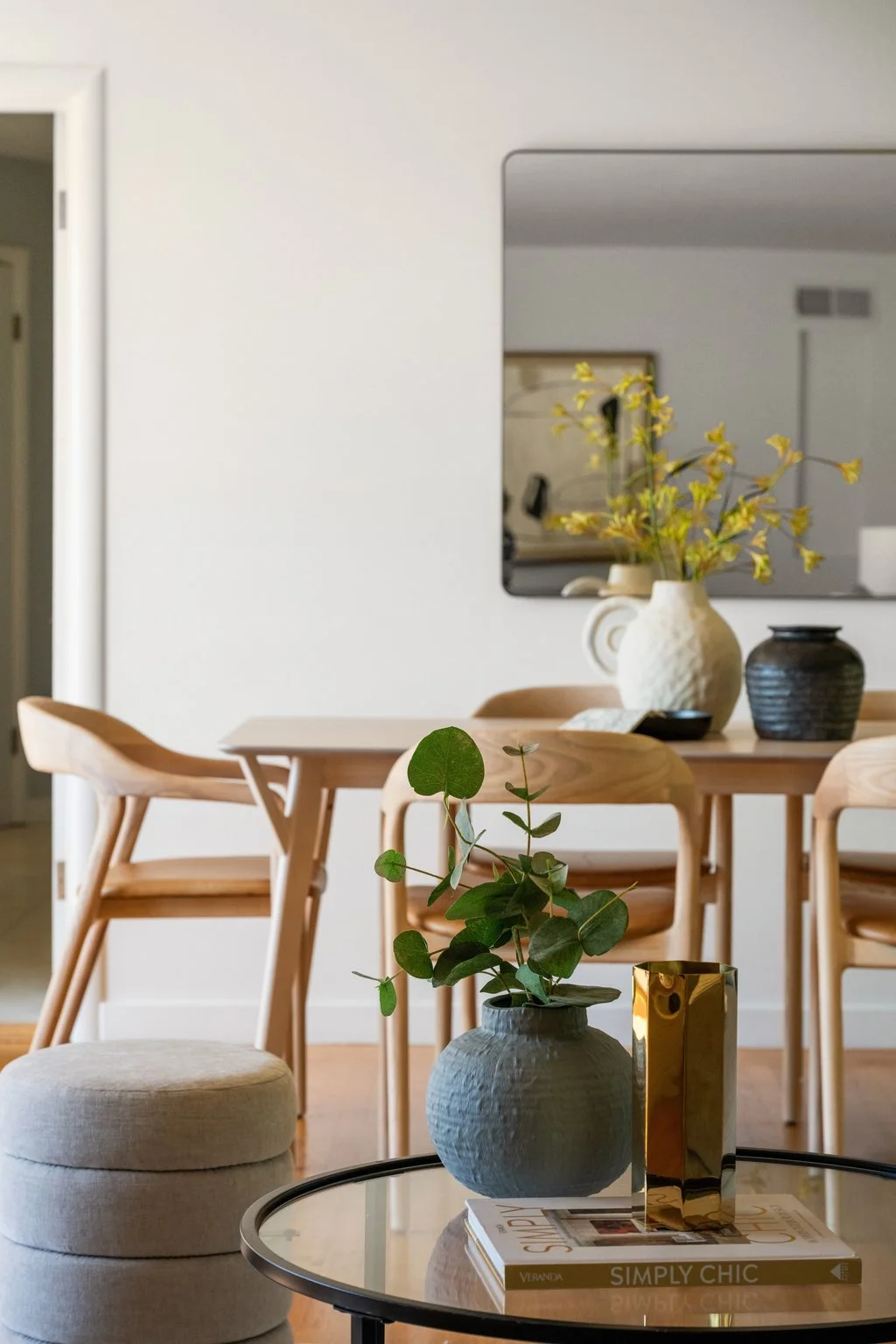Interior of a modern dining area with a wooden table, chairs, and decorative vases with plants. A mirror on the wall reflects part of the room.