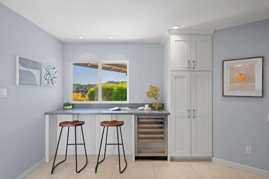 Modern kitchen with white cabinetry, light gray walls, two brown bar stools, a window revealing greenery, a small wine fridge, and framed artwork.
