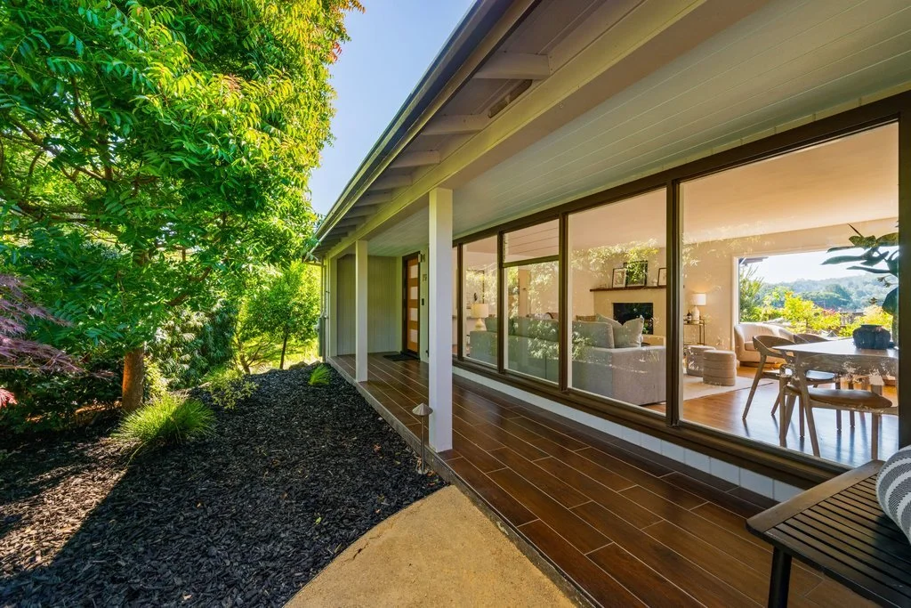 View of a modern house with large glass windows overlooking a lush garden with trees, plants, and mulch.
