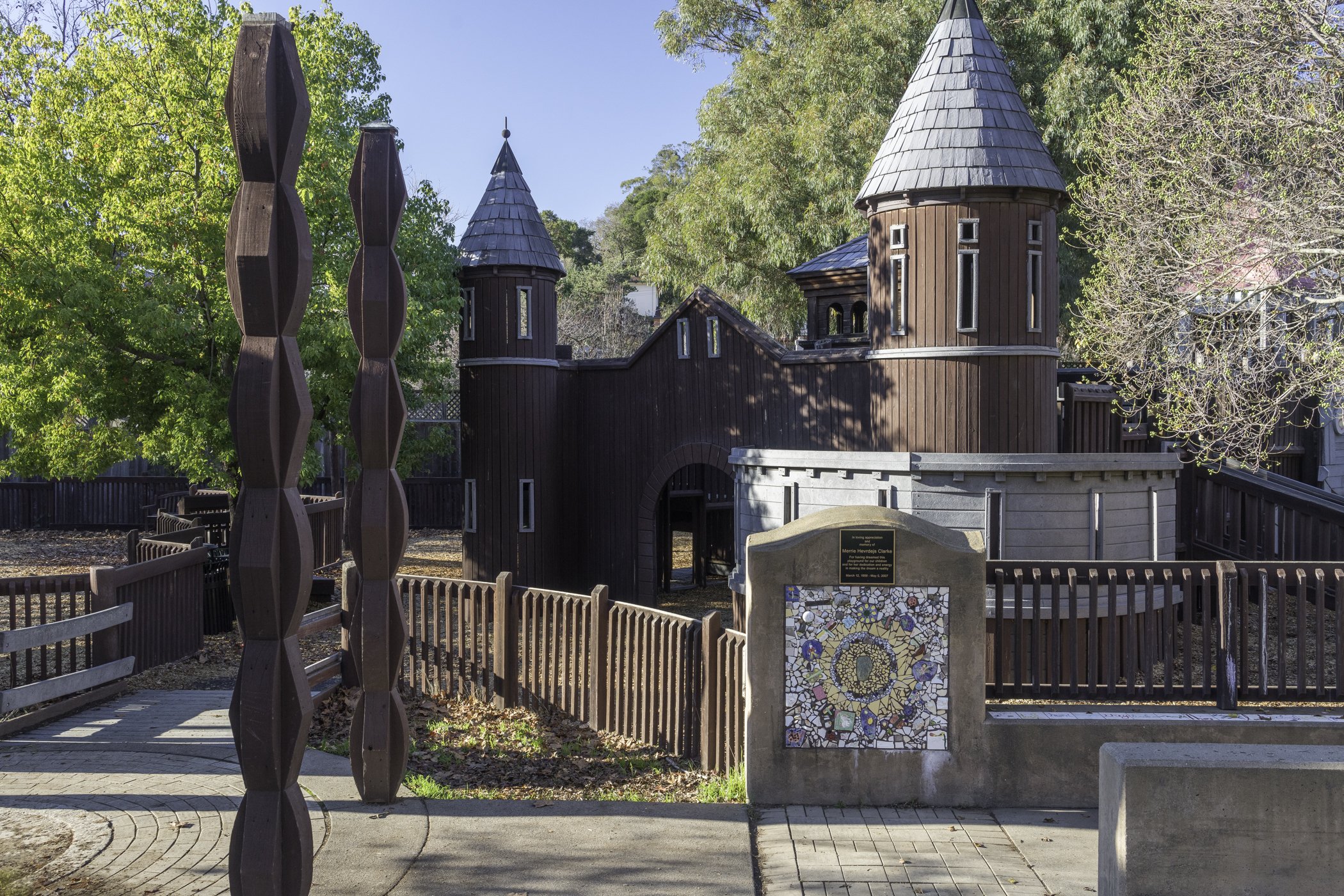 A children's play area featuring a castle-like play structure with towers and a wooden fence surrounding it. There is an abstract sculpture made of wooden pieces in the foreground and trees with green and yellow leaves in the background.