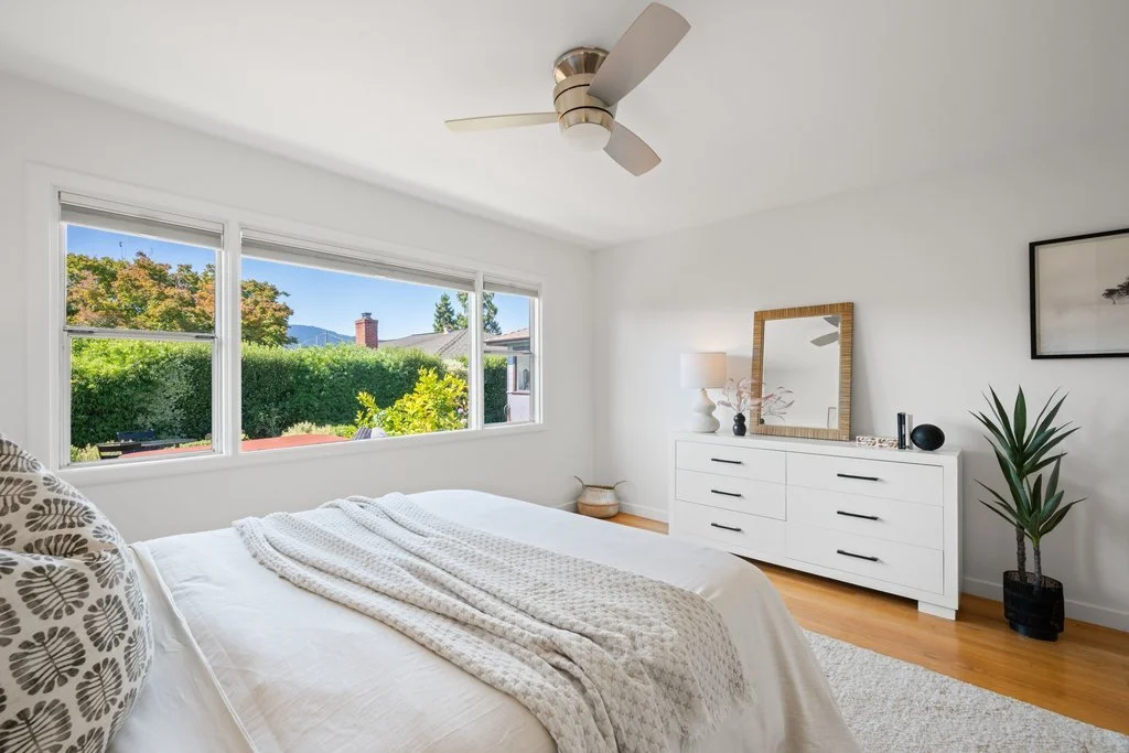 Bright bedroom with large window, white bed, decorative pillows, white dresser with mirror, potted plant, and ceiling fan.