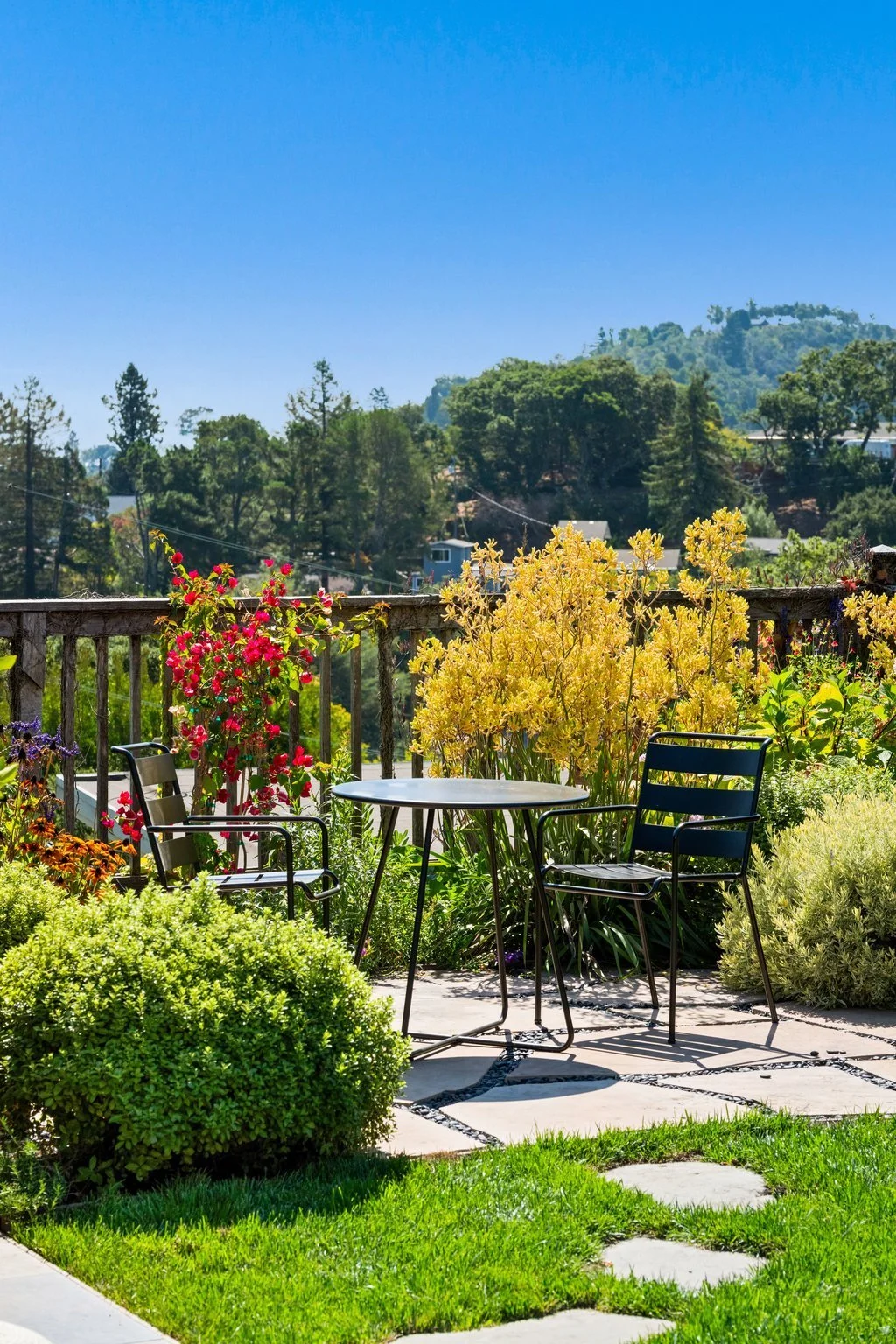 A backyard patio with a round table and two black chairs surrounded by colorful flowers and green bushes, overlooking a wooded hillside under a clear blue sky.